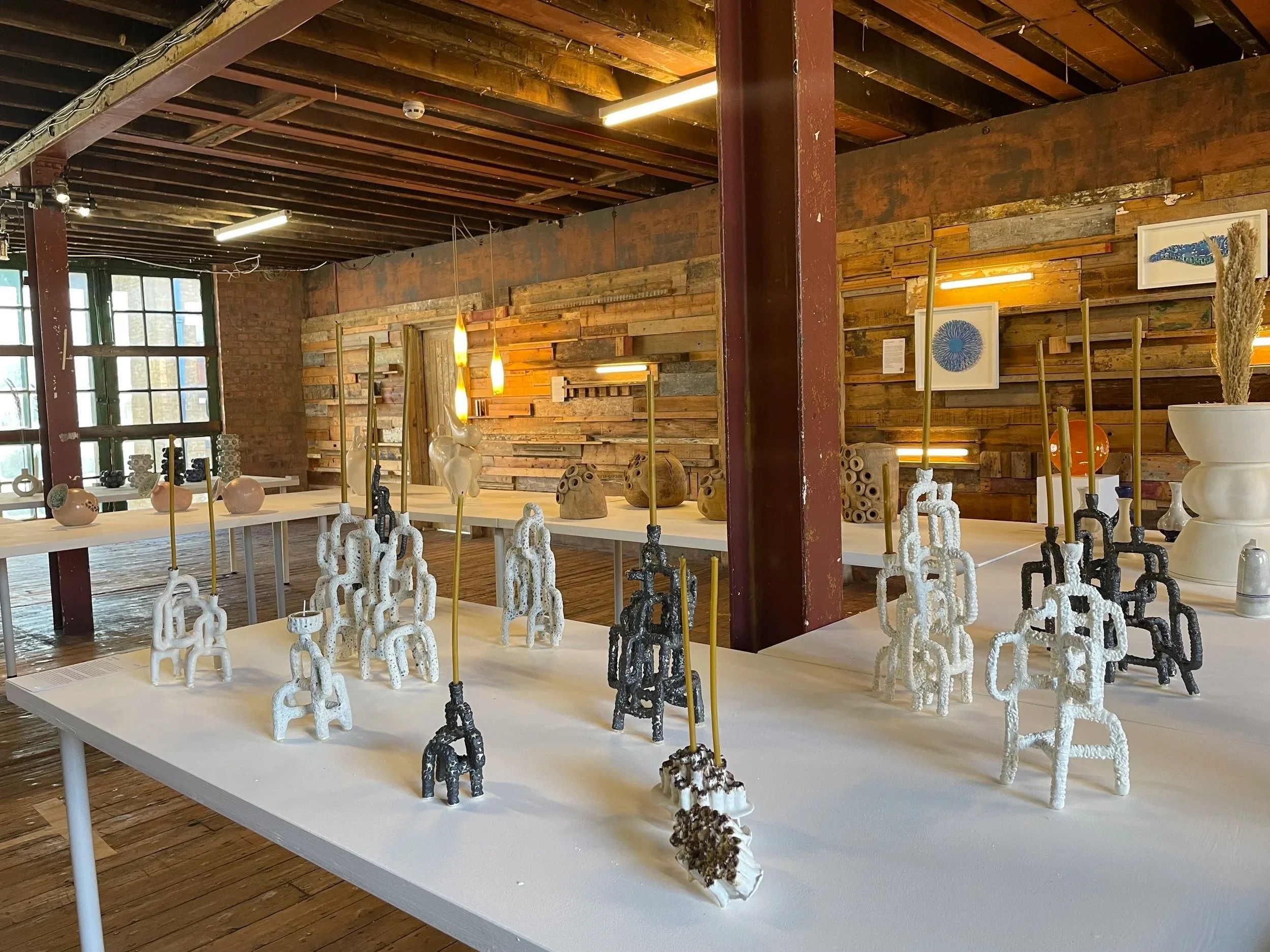 Interior of an art gallery with wooden walls and ceiling, displaying ceramic and sculpture art on white tables, illuminated by hanging and wall lighting.