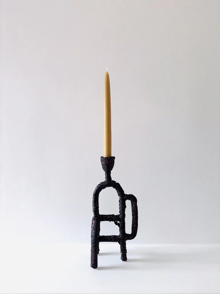 A black and terracotta decorative candlestick holding a yellow beeswax taper candle, against a plain white background.