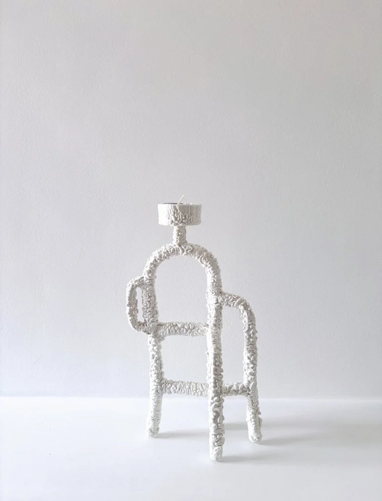 A white decorative tall tealight holder with a small white candle on top, placed on a white surface against a plain white wall.