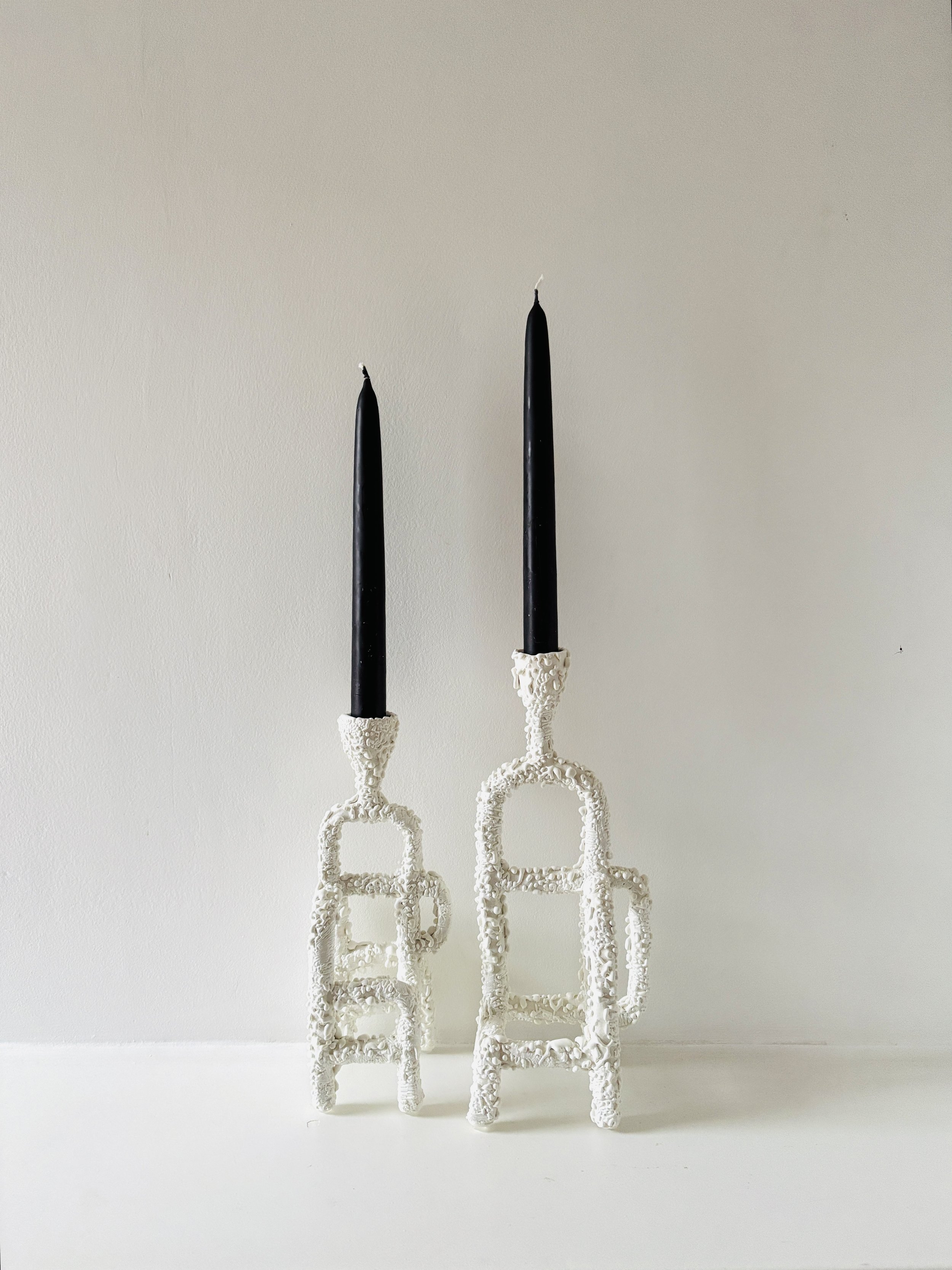 Two black candles in white ornate holders. The holder of the right candle is larger and more elaborate than the left.
