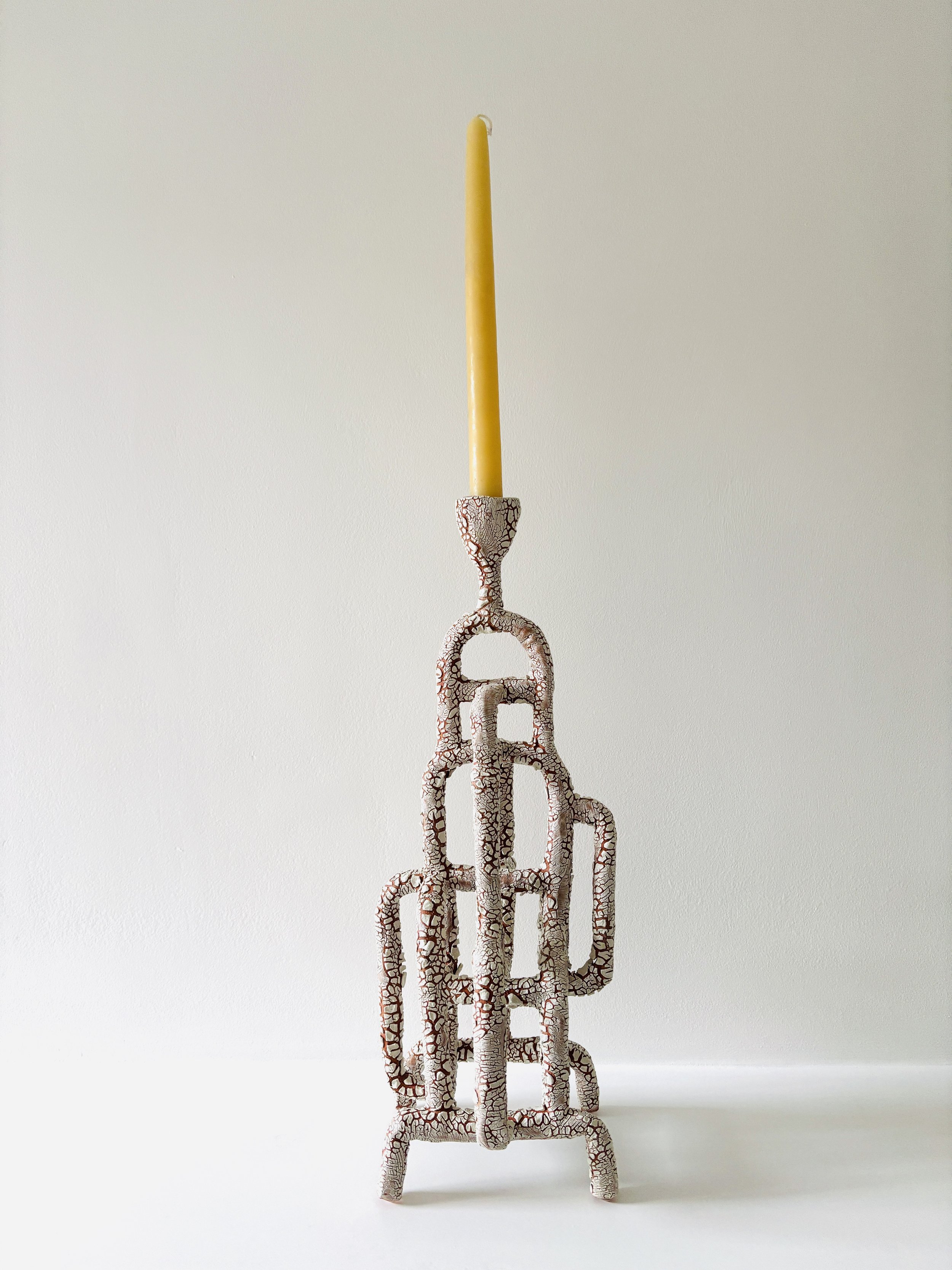 A ceramic sculpture resembling a tall, thin sculpture with multiple intertwined legs, topped with a long, upright candle, set against a plain white background.