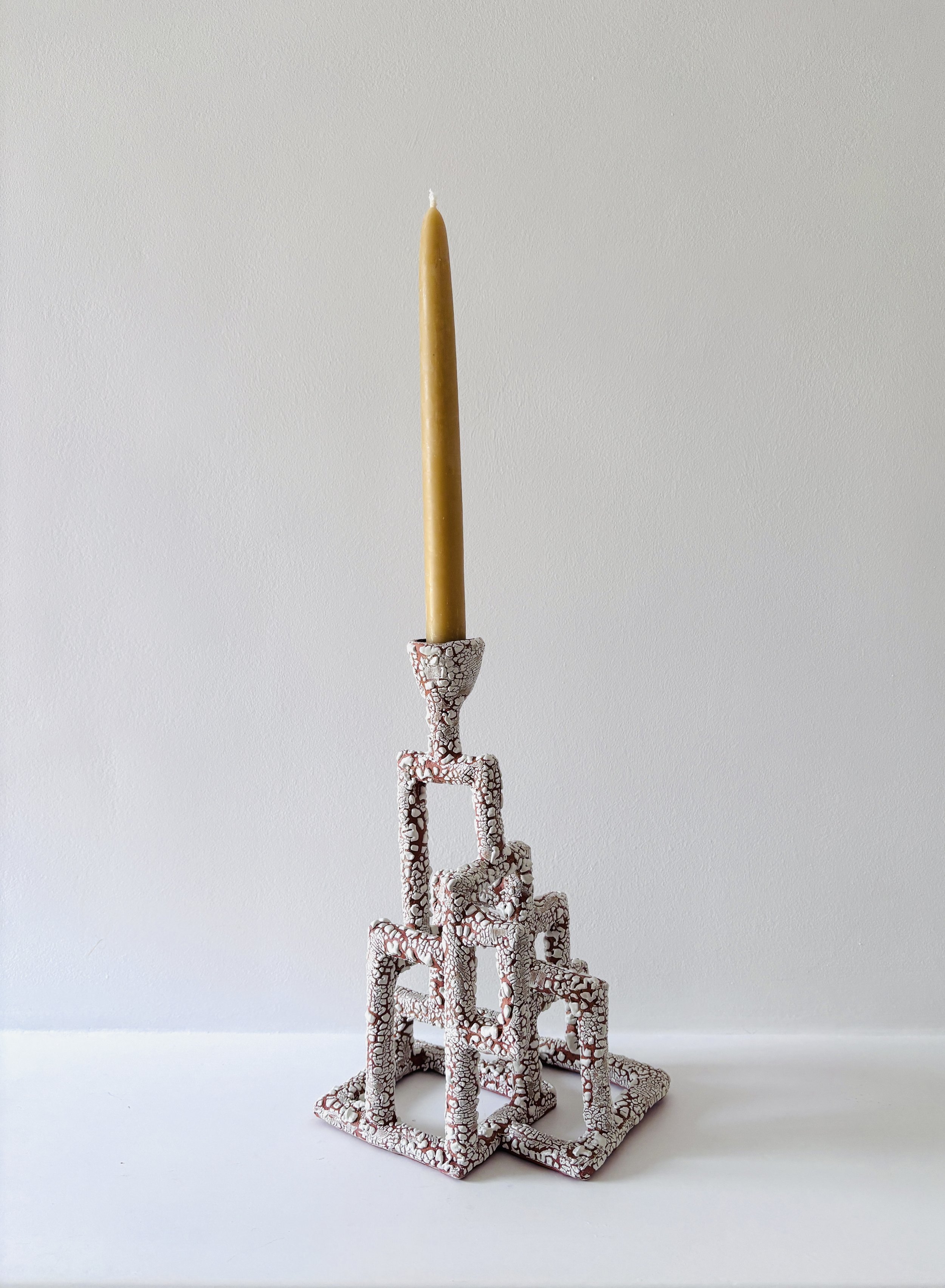 A tall, yellow beeswax candle is placed on top of a tangled ceramic sculpture with a cracked white and terracotta glaze pattern, set against a plain light gray background.
