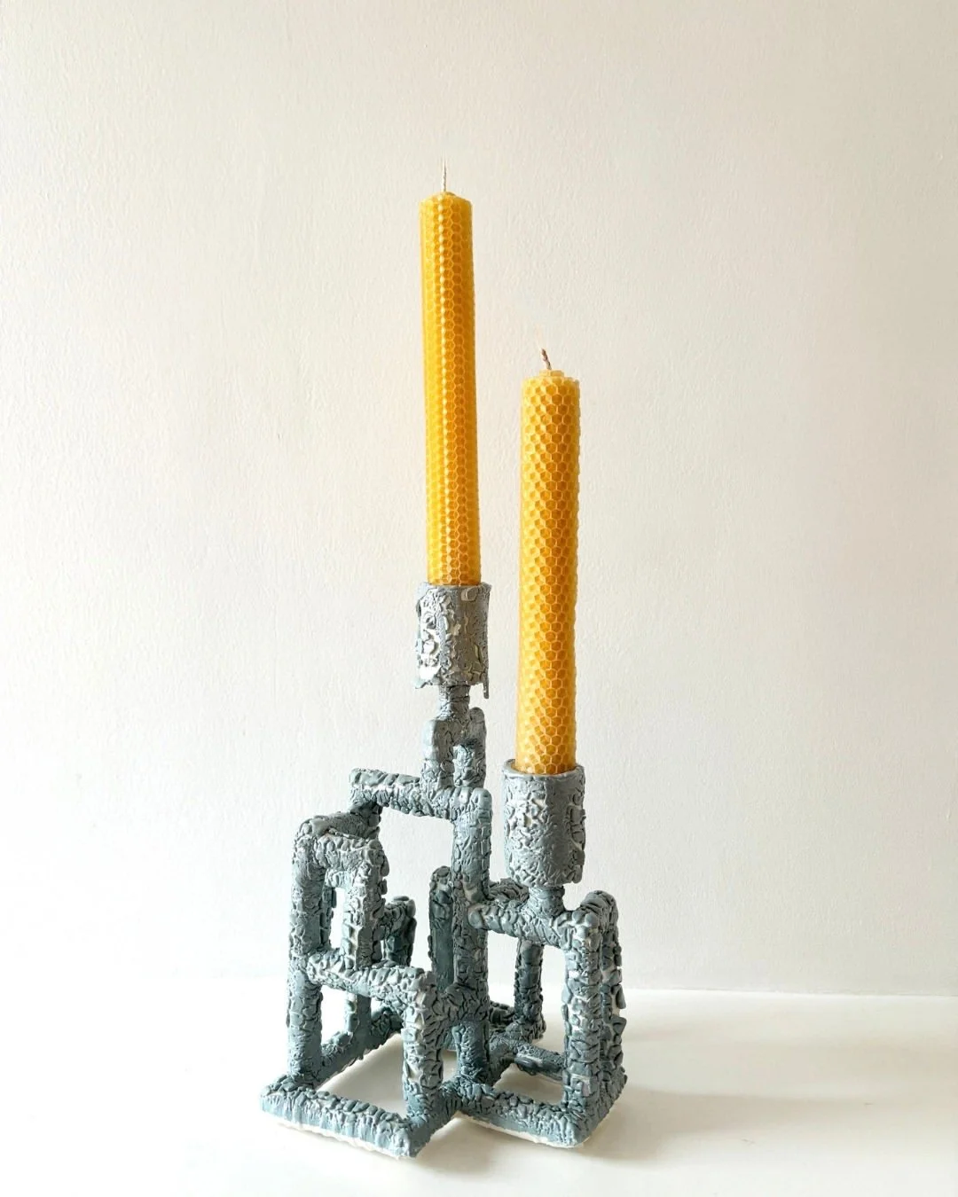 Two yellow beeswax candles with honeycomb pattern sit in sea storm , textured, candle holders on a white surface against a plain white background.