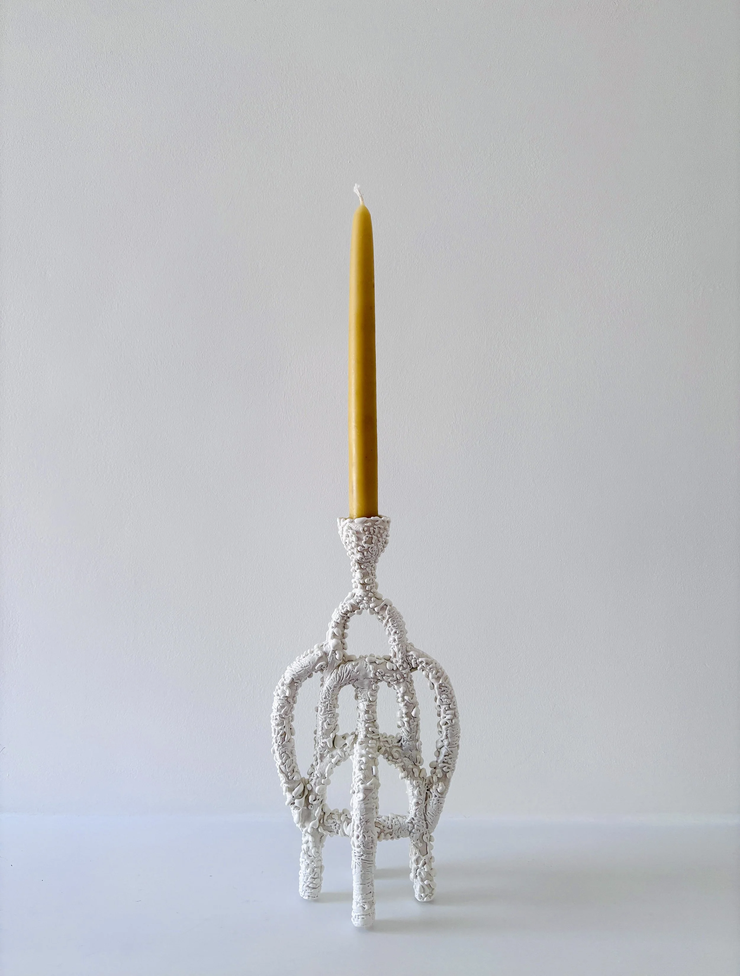 A tall, yellow beeswax candle standing upright in a white, ornate candle holder shaped like a tree trunk with branches.