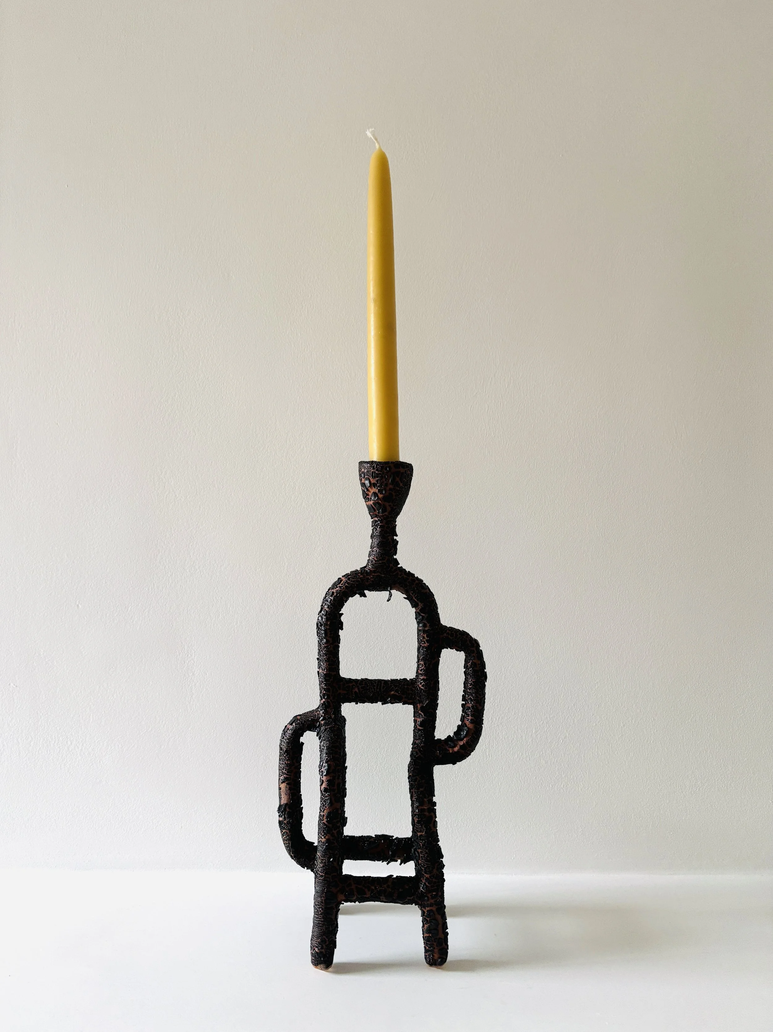 A tall yellow candle held in a black, ornate holder against a plain white wall.