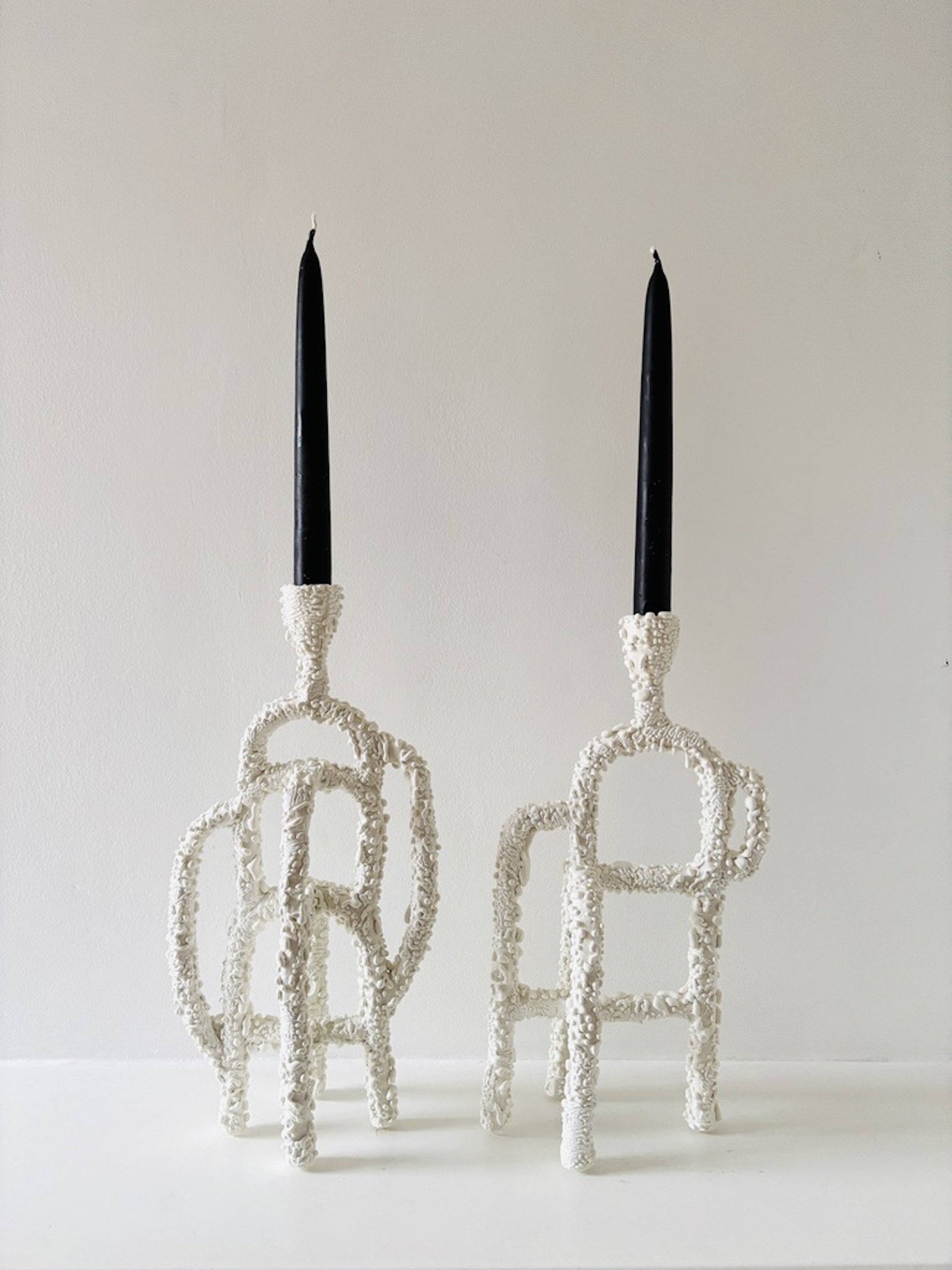 Two black candles in white, textured candle holders arranged in a row against a plain white background.