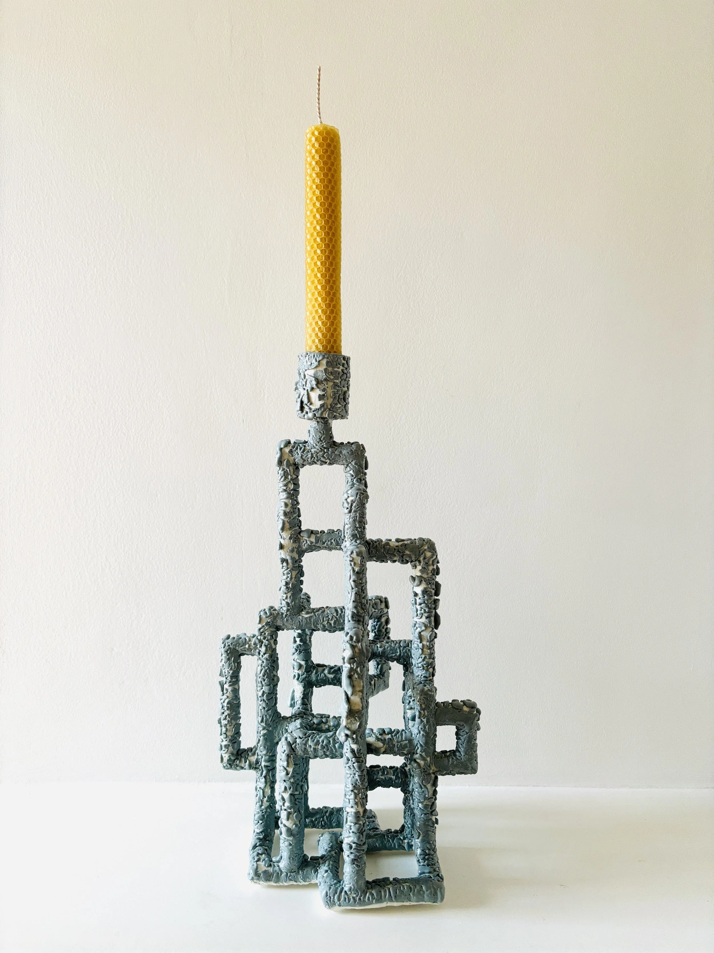 A tall, pale yellow, textured candle placed on a decorative ceramic holder with textured glaze in a grey green colour