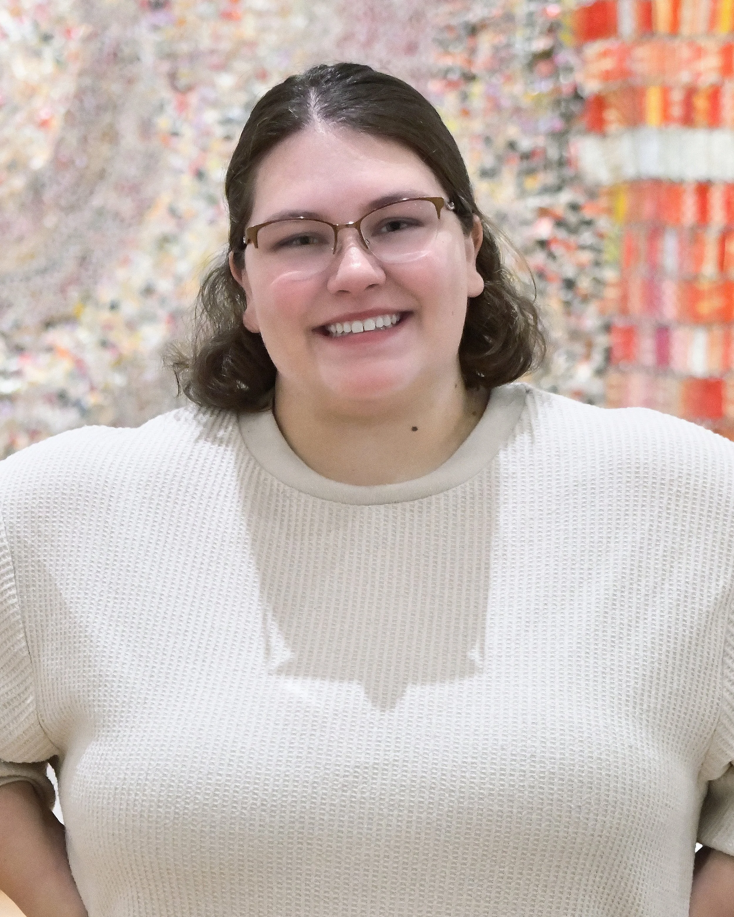 A woman with glasses and brown hair smiling, wearing a cream sweater, standing in front of a colorful wall.
