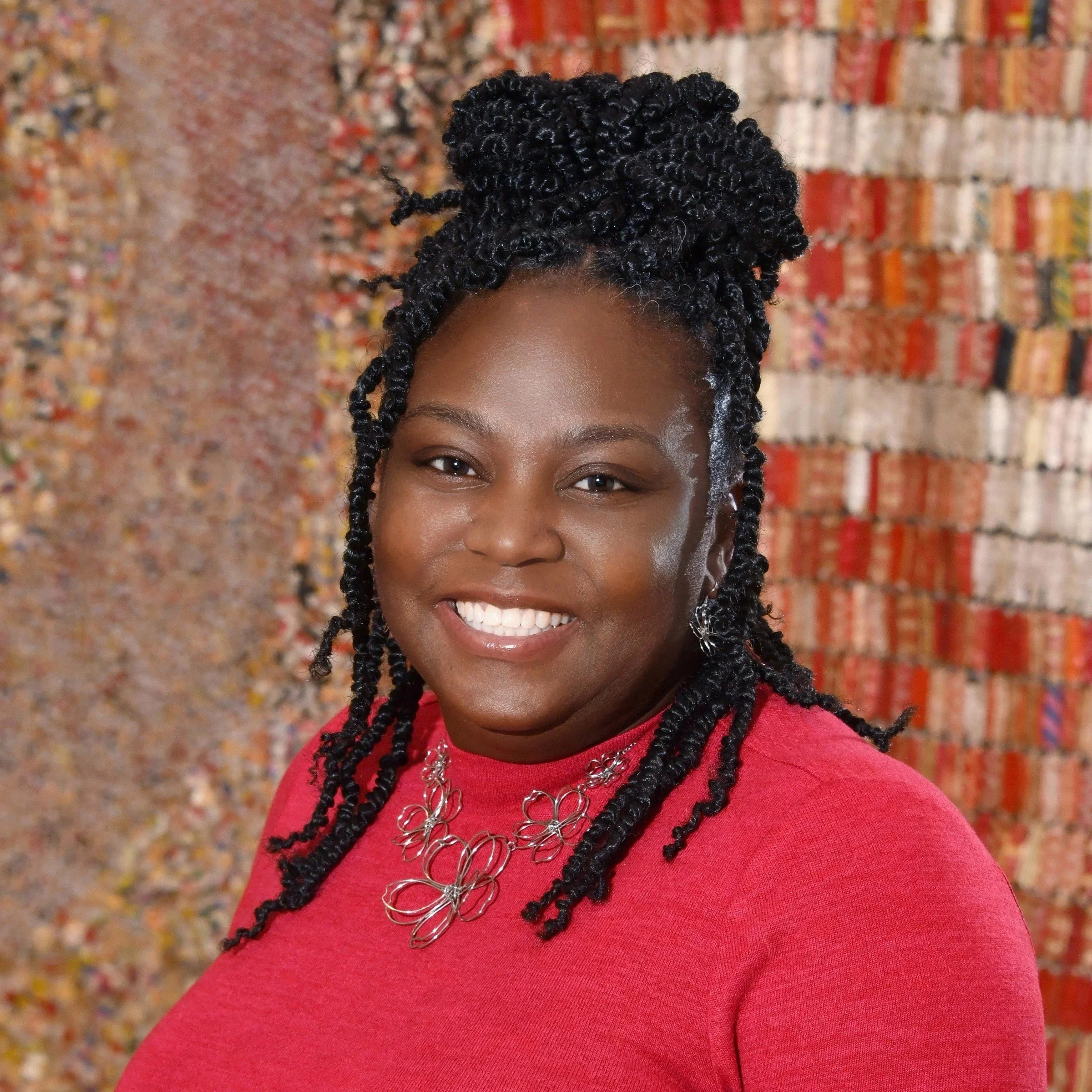 Portrait of a smiling woman with black braided hair, wearing a red top and silver jewelry, standing in front of a background with patterned textiles.