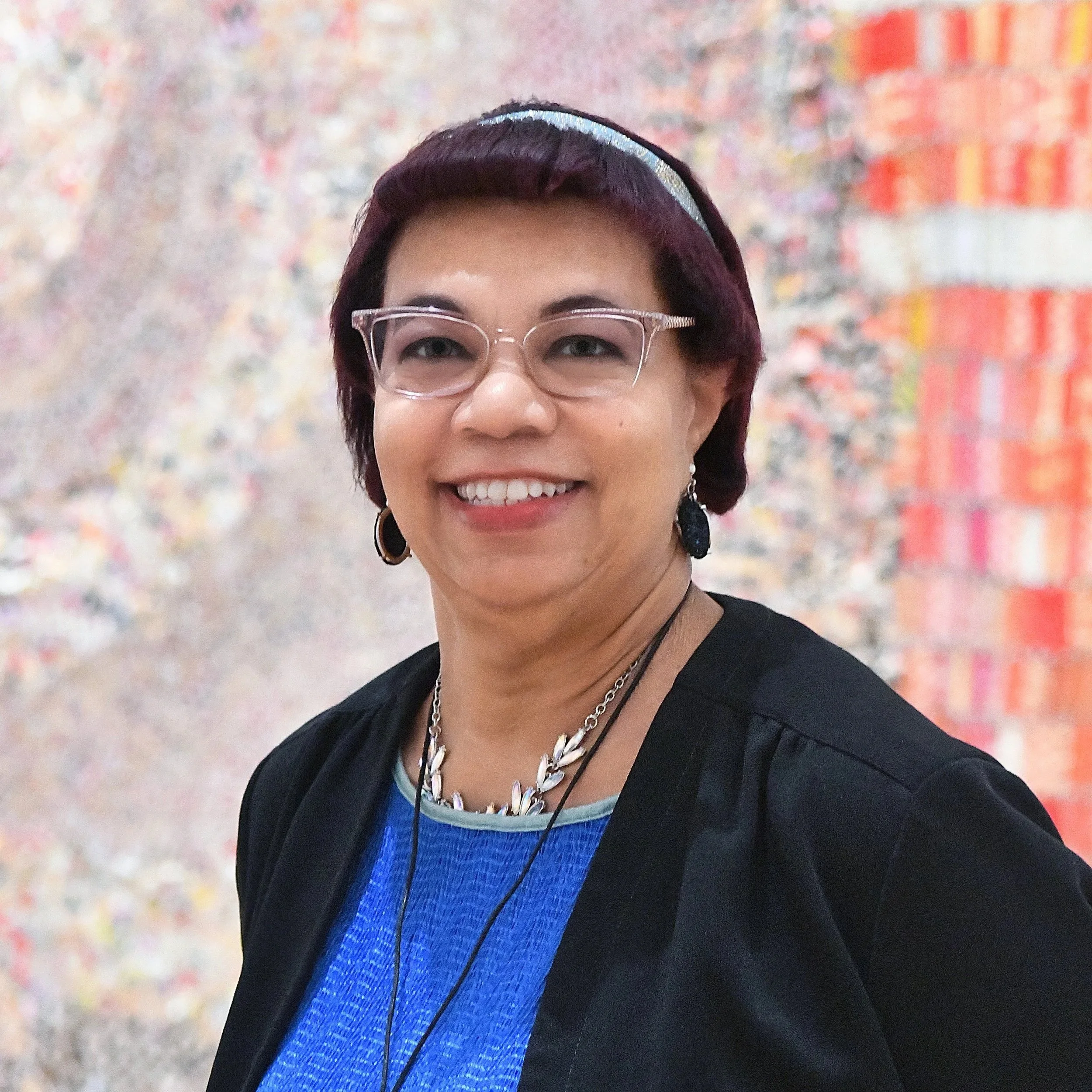 A woman with short hair, wearing clear glasses, black earrings, a blue top, black jacket, and jewelry, smiling in front of a colorful textured background.