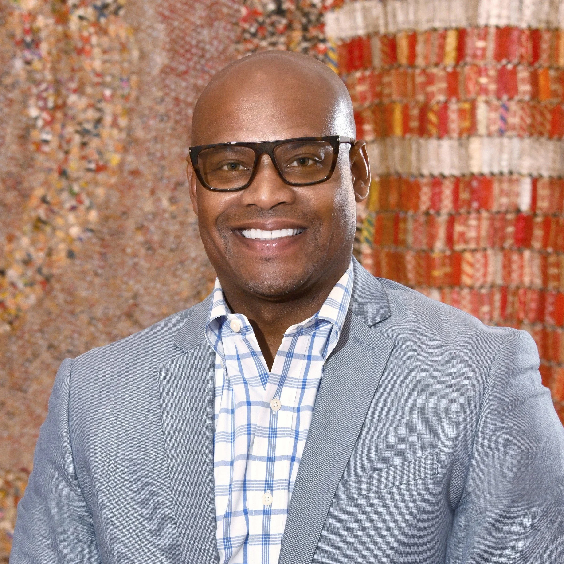 A smiling man wearing glasses, a gray suit, and a blue plaid shirt standing in front of an abstract, colorful textured background.
