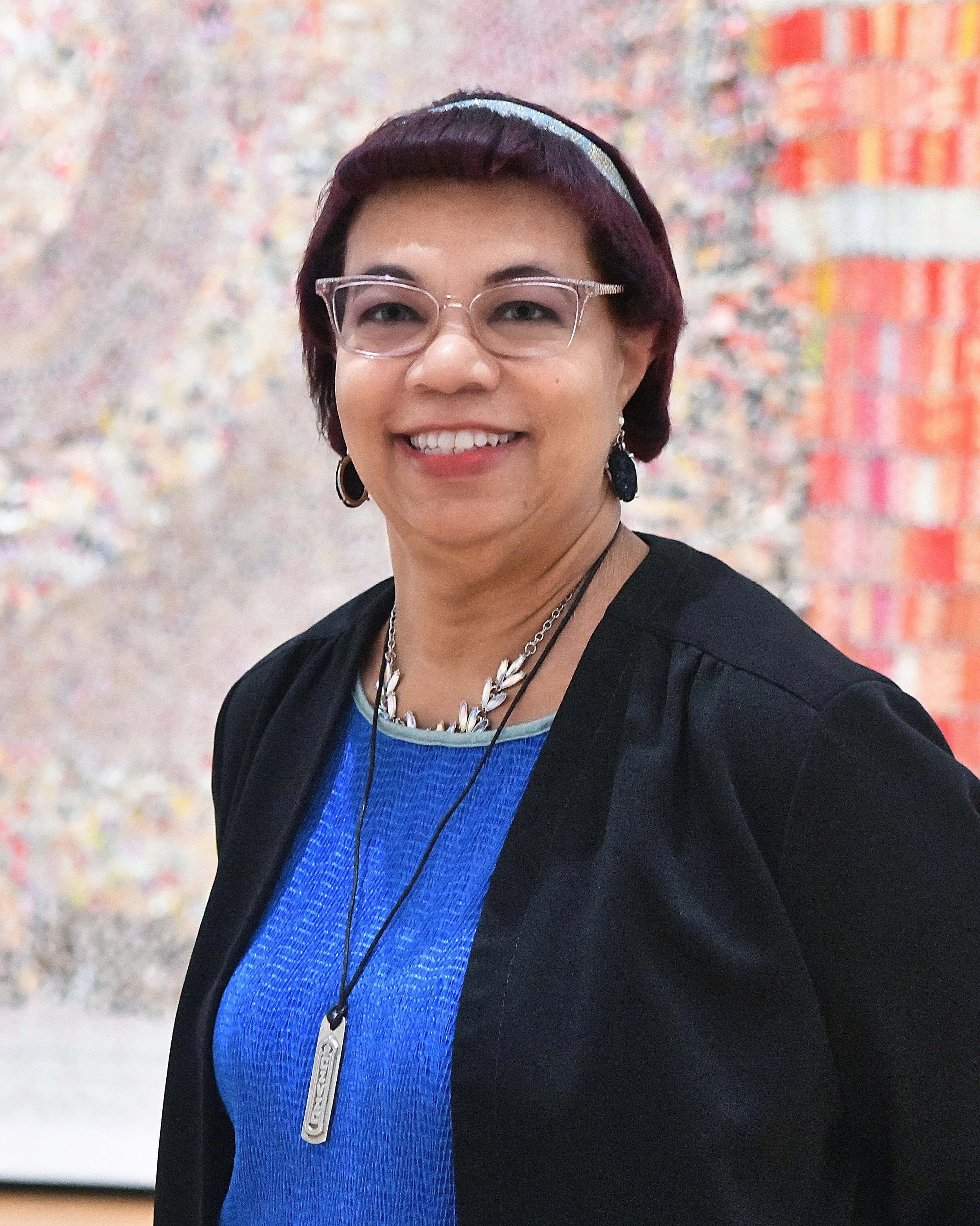 A woman with short dark hair, wearing glasses, a black jacket over a blue top, and jewelry, standing in front of a colorful abstract background.