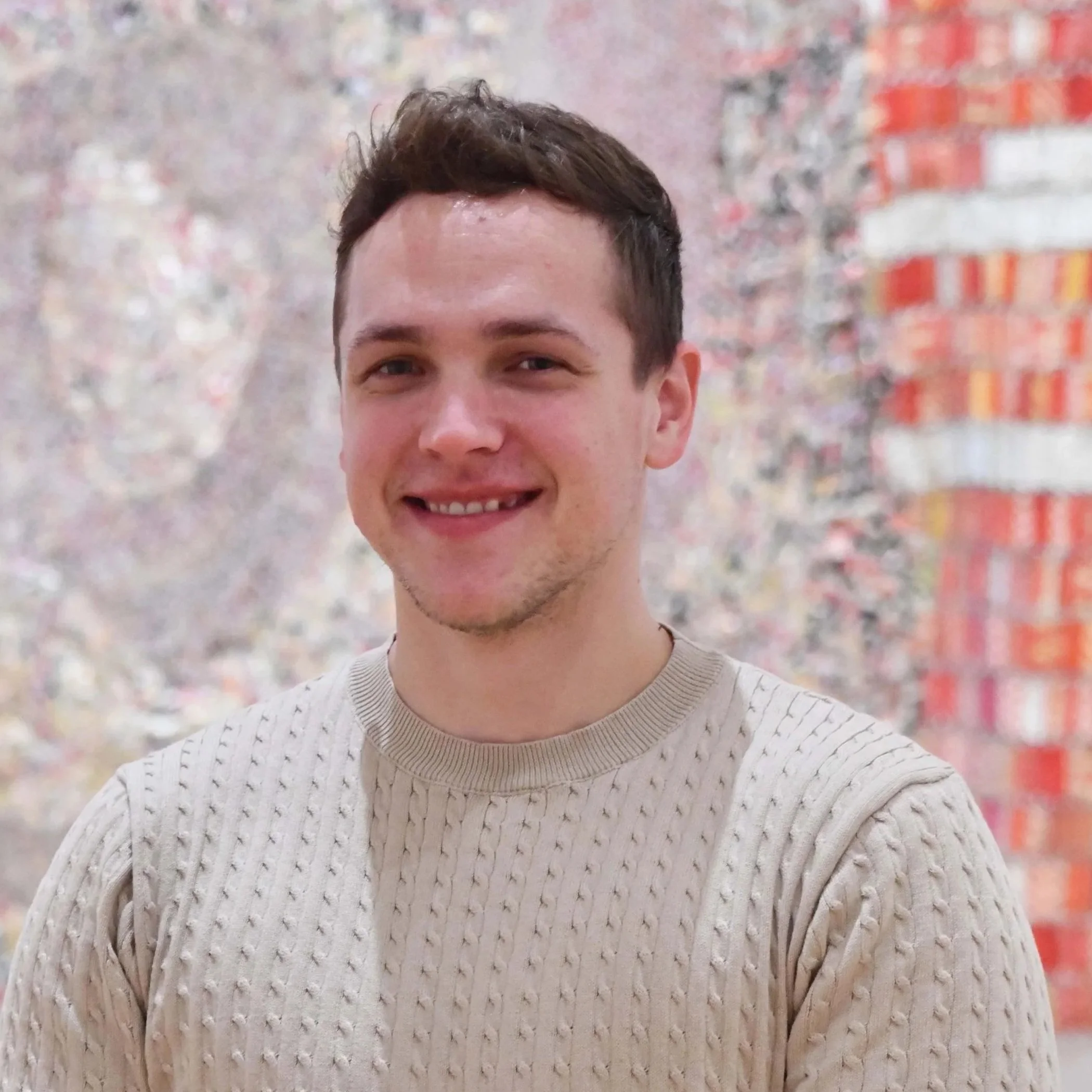 Young man with short brown hair smiling, wearing a beige cable-knit sweater, standing outdoors with blurred trees and a building in the background.
