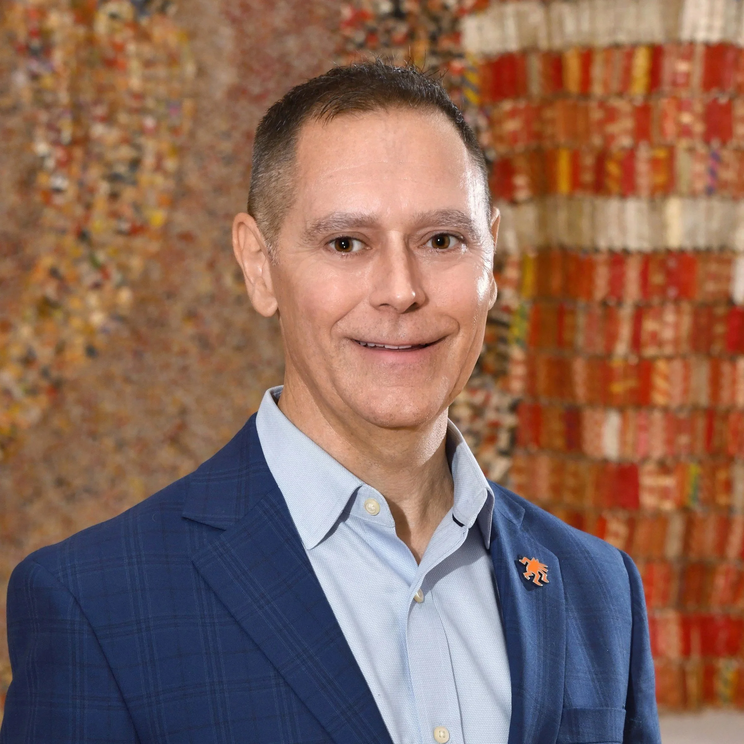 A man in a blue checkered suit and light blue shirt standing in front of a colorful, abstract tapestry.