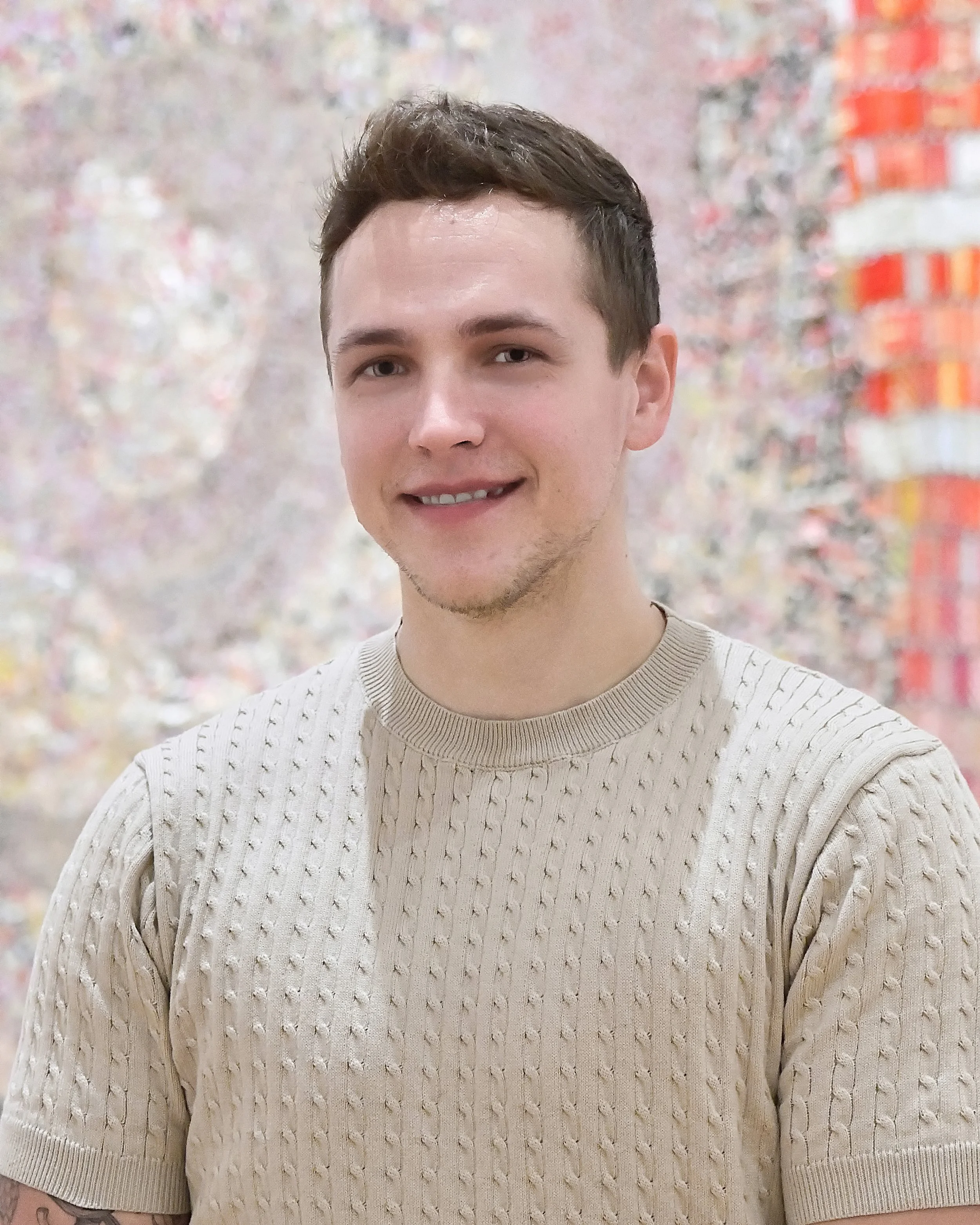 A young man with short brown hair, fair skin, and light facial hair. He is standing in the Akron Art Museum in front of a colorful, blurry background. He is smiling slightly and wearing a beige knitted sweater.