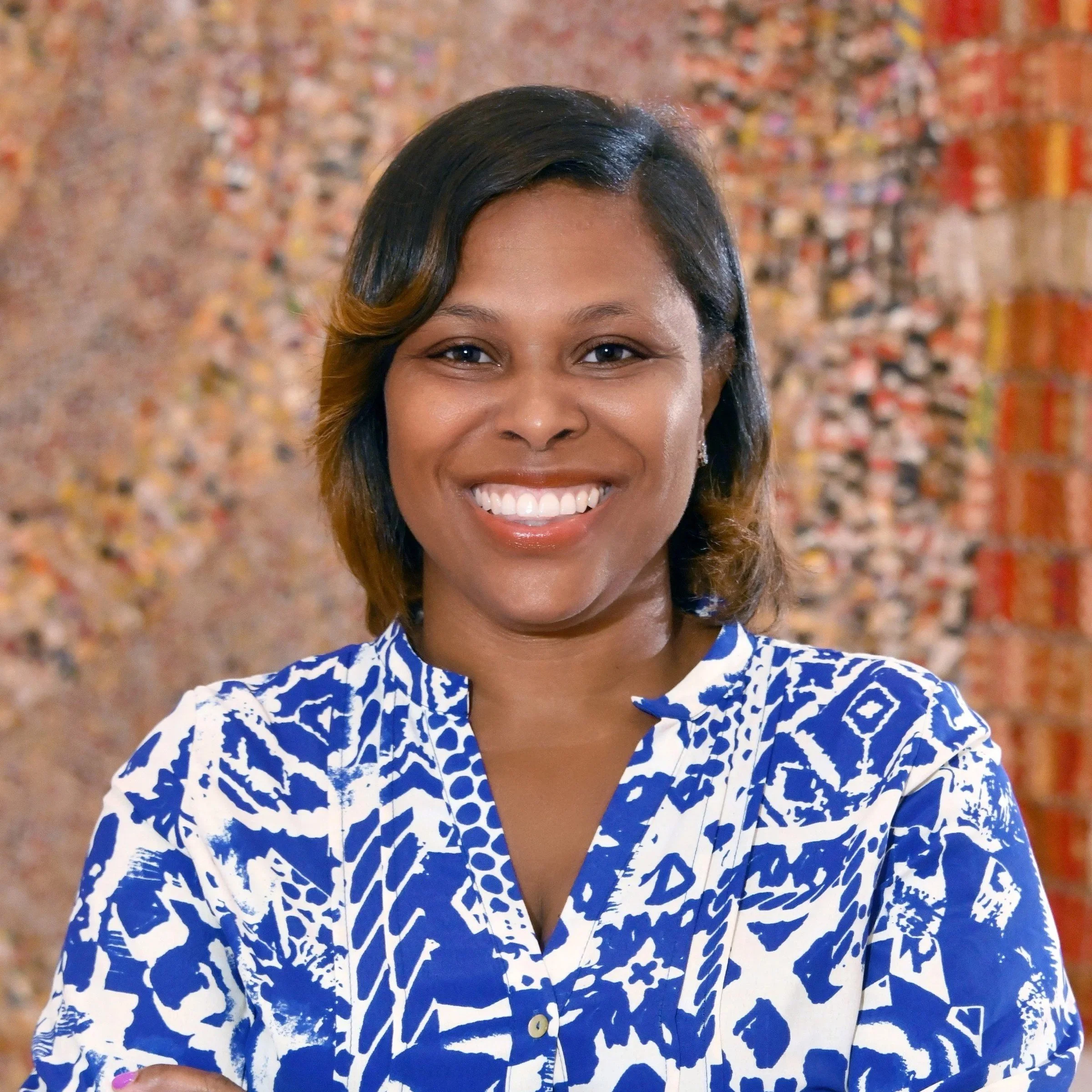 Smiling woman with shoulder-length black hair and light brown highlights, wearing a blue and white patterned blouse, standing in front of a colorful textured wall.