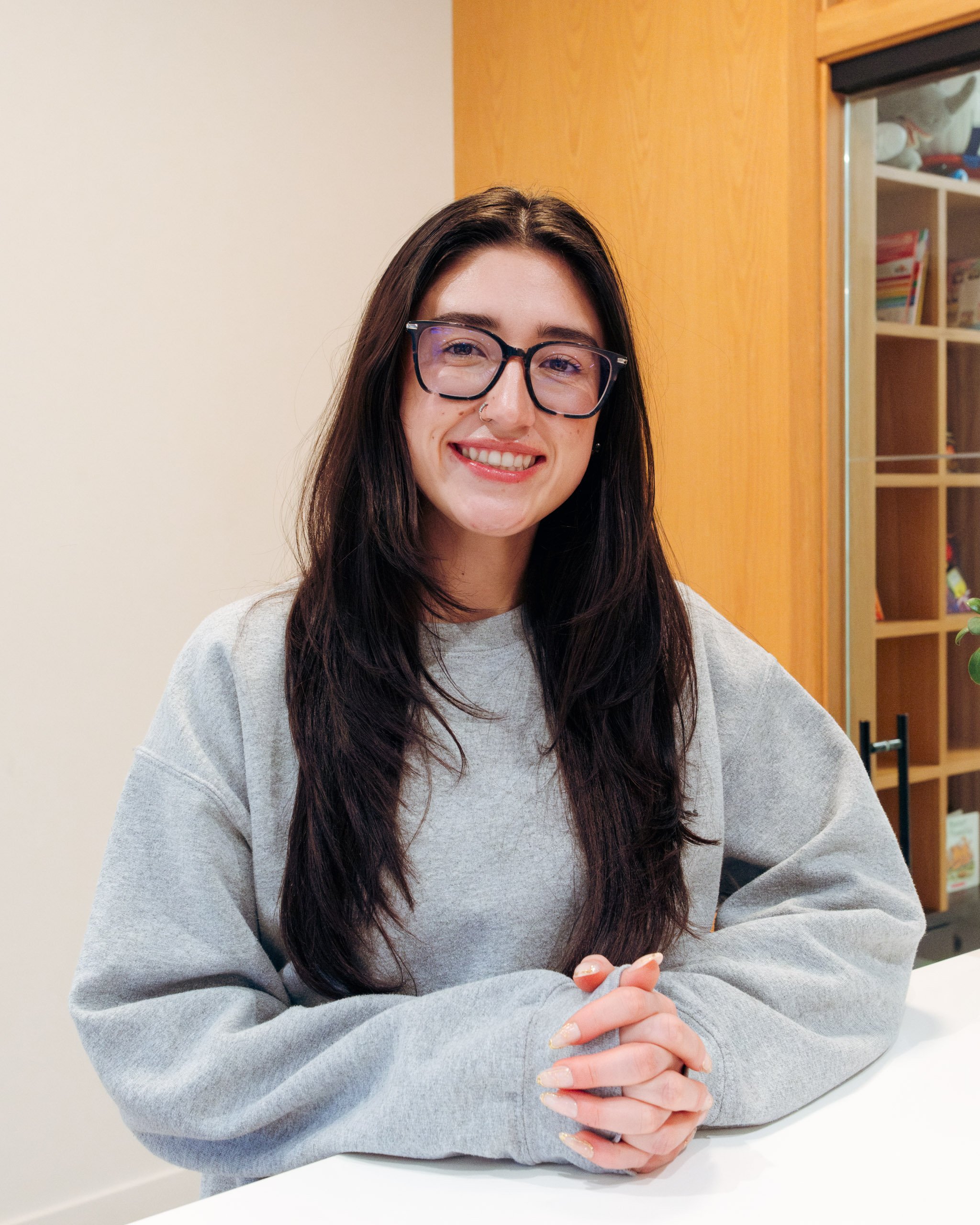 A woman in a navy blue zip-up jacket smiles at the camera for a portrait. She is wearing glasses and has long brown hair that has been parted in the middle.
