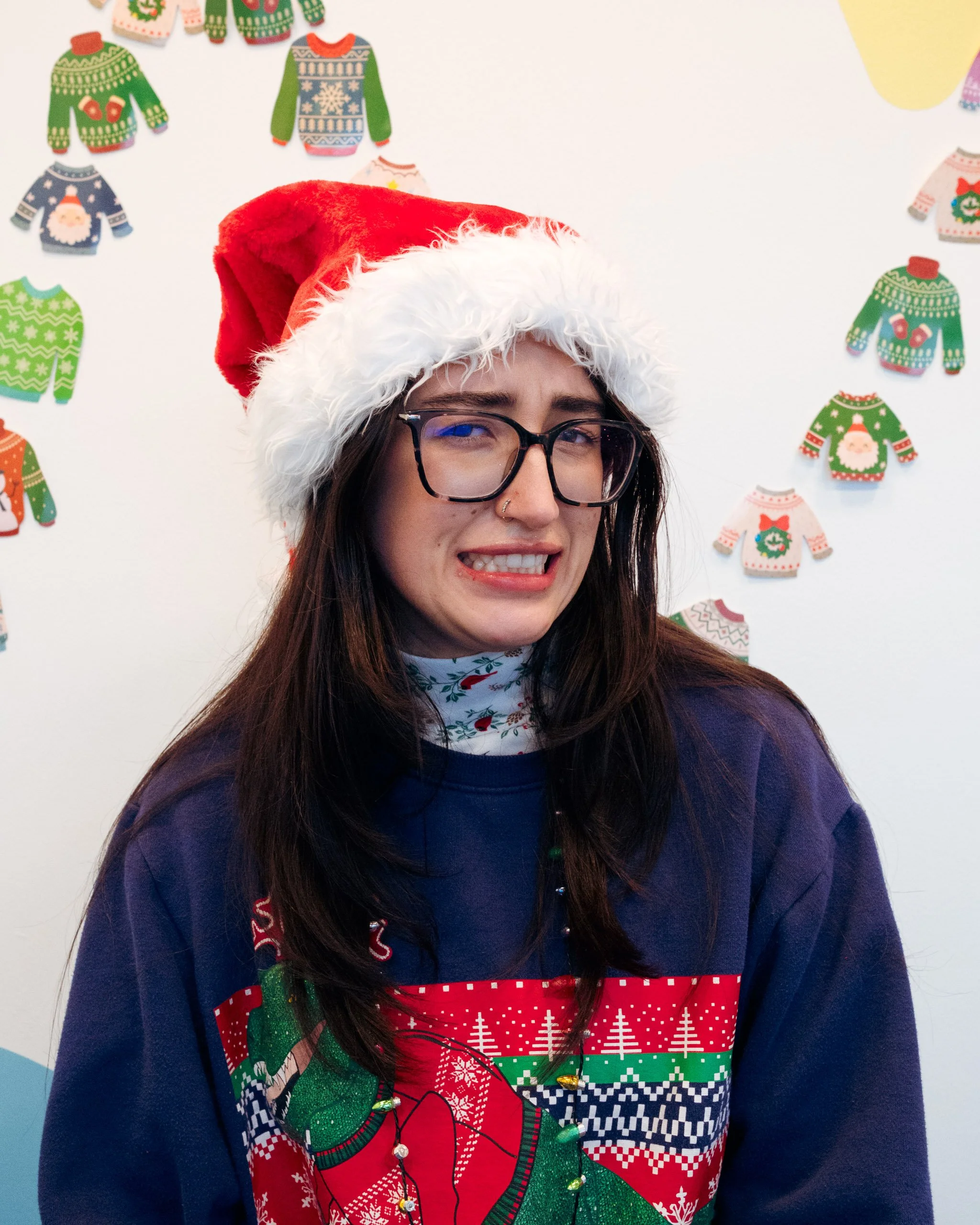 A woman in a navy blue zip-up jacket smiles at the camera for a portrait. She is wearing glasses and has long brown hair that has been parted in the middle.