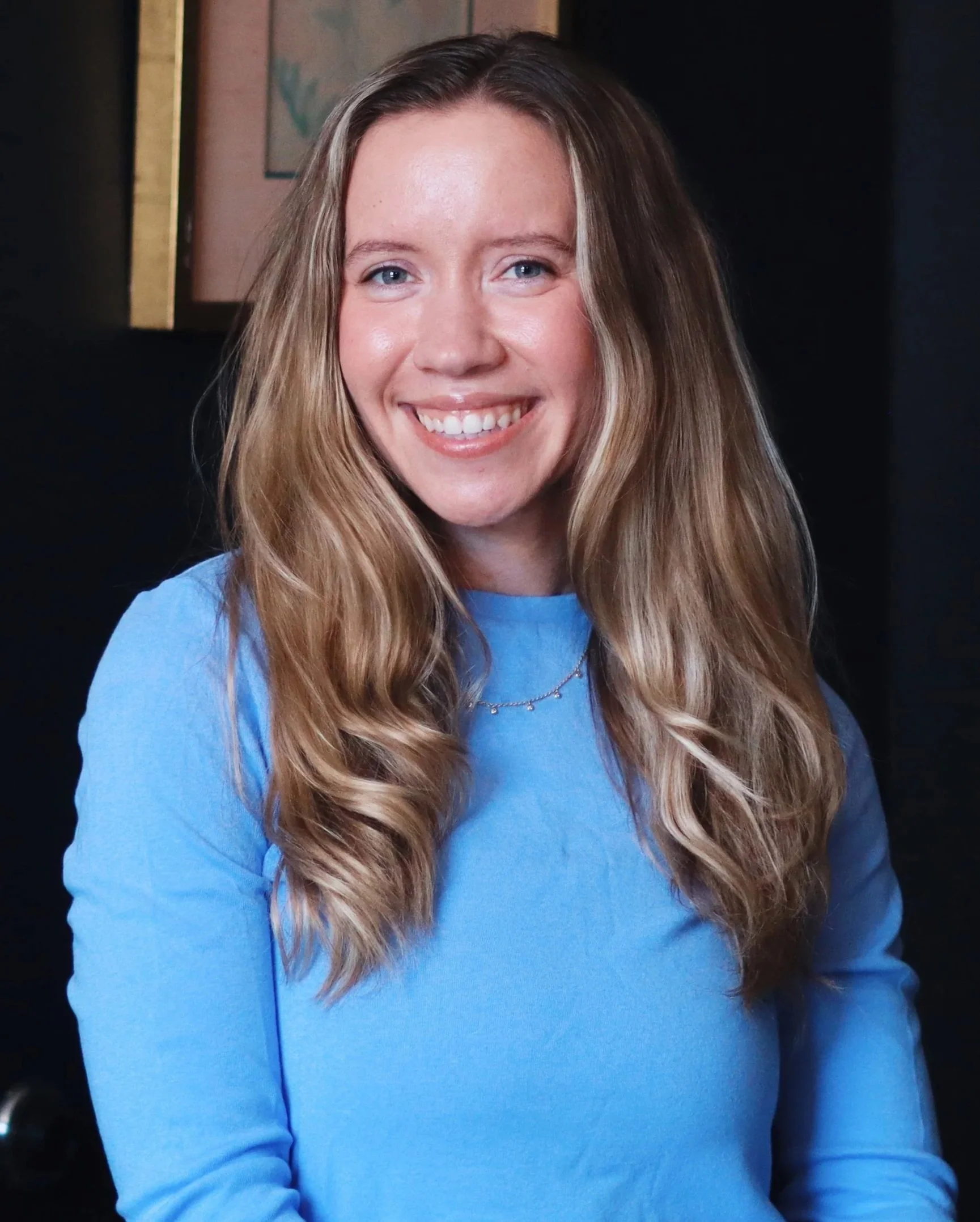 Close-up of a smiling woman with long, wavy blonde hair, wearing a blue top and a silver necklace, inside a voice studio with a dim background and framed artwork.