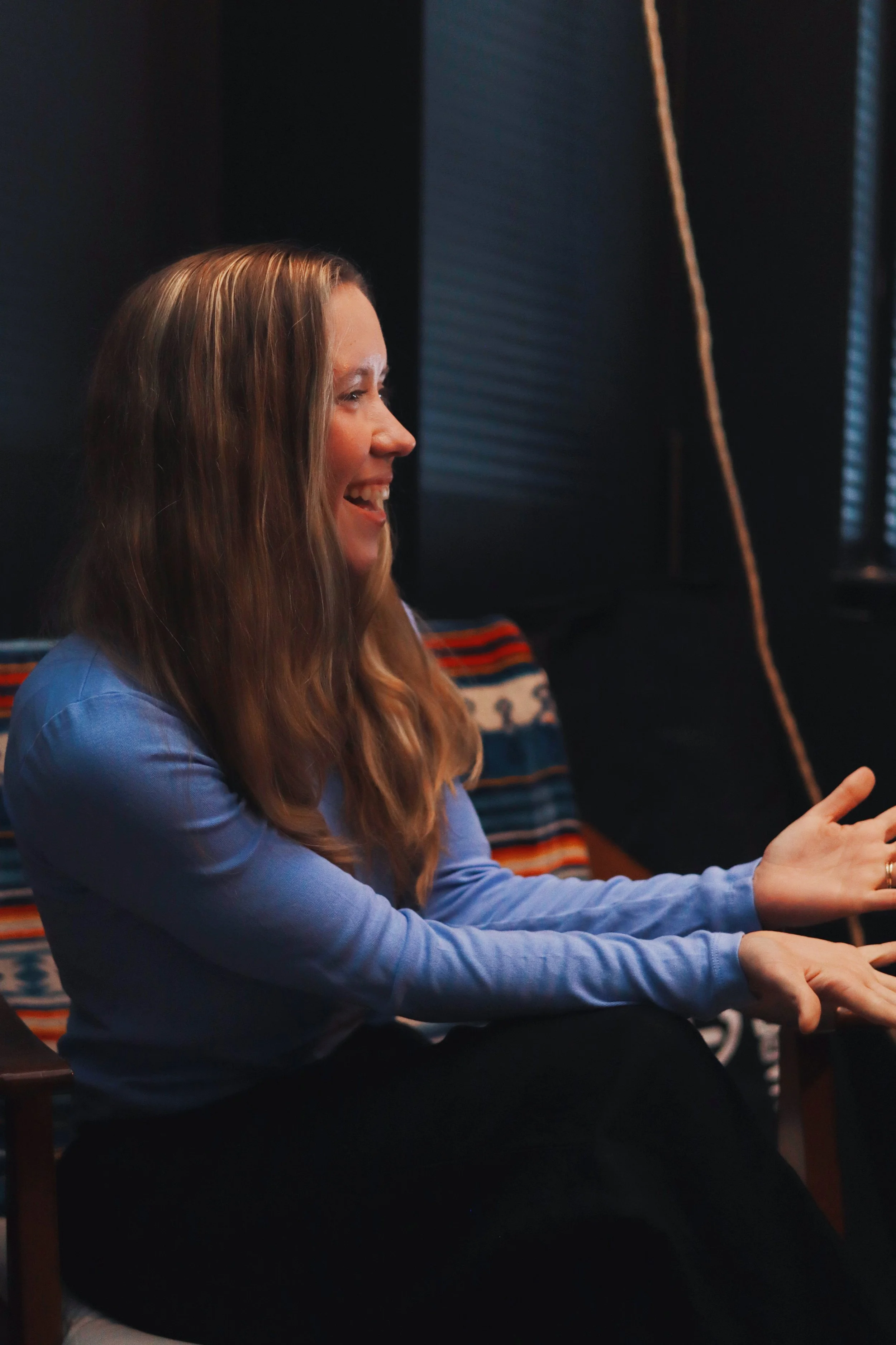 A voice teacher with long reddish-blonde hair smiling and having a conversation about singing, with part of a patterned textile visible behind her.