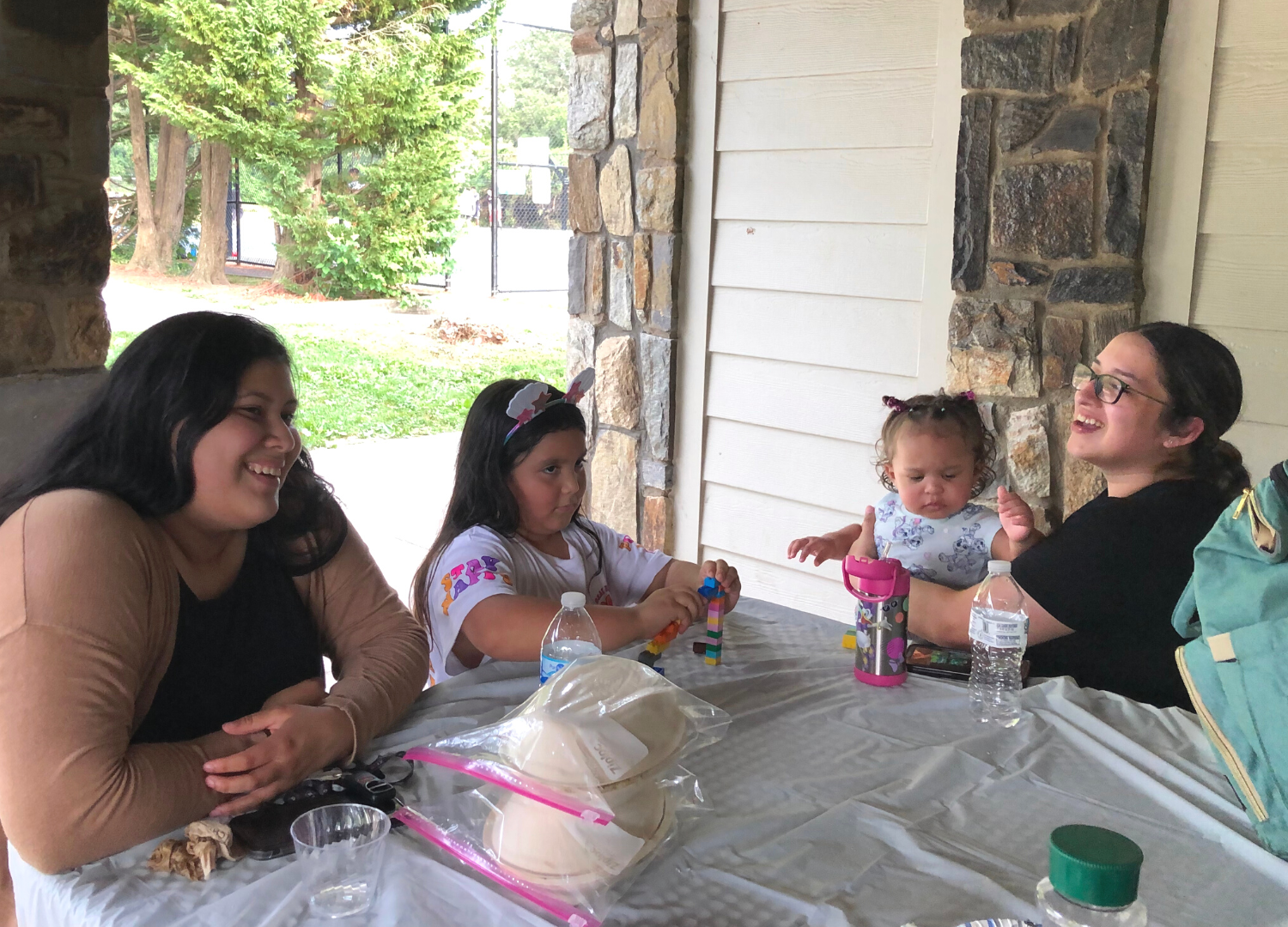 Two women, Generation Hope Scholars, laughing and sitting with their daughters at an outdoor table at a park.