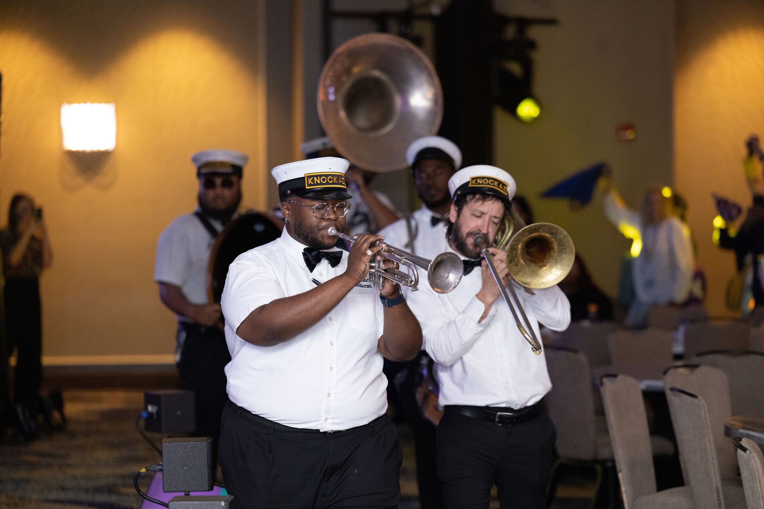 A marching band performing indoors, with musicians playing trumpet, trombone, and other brass instruments. They wear white shirts, black bow ties, and hats labeled "Knockaz."