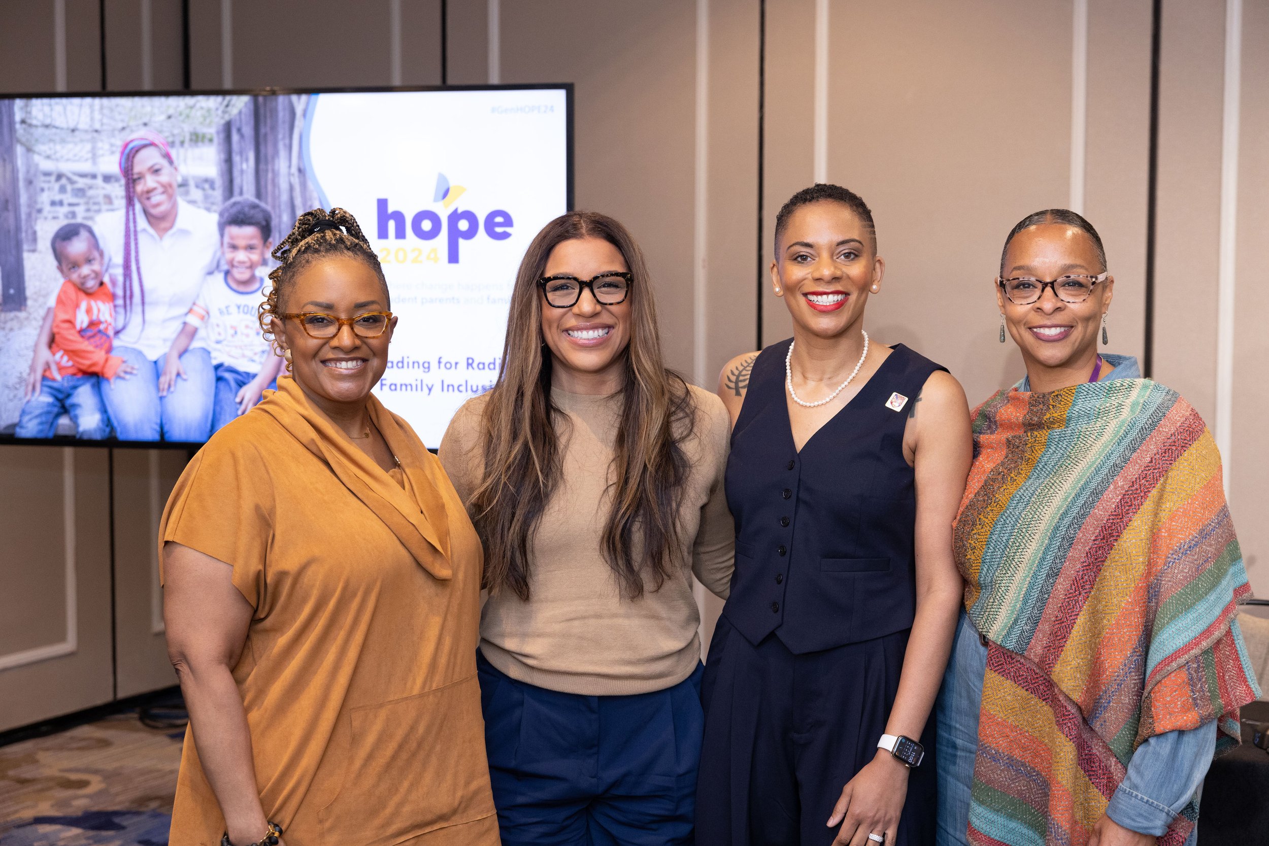 Four women smiling at a "Hope 2024" event, with a screen displaying the event logo and an image of a woman with two children in the background.