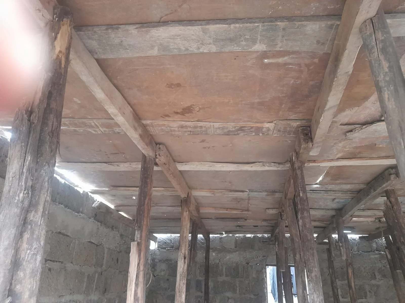 Interior construction site with wooden beams and plywood ceiling, supported by vertical wooden posts. The walls are made of concrete blocks.