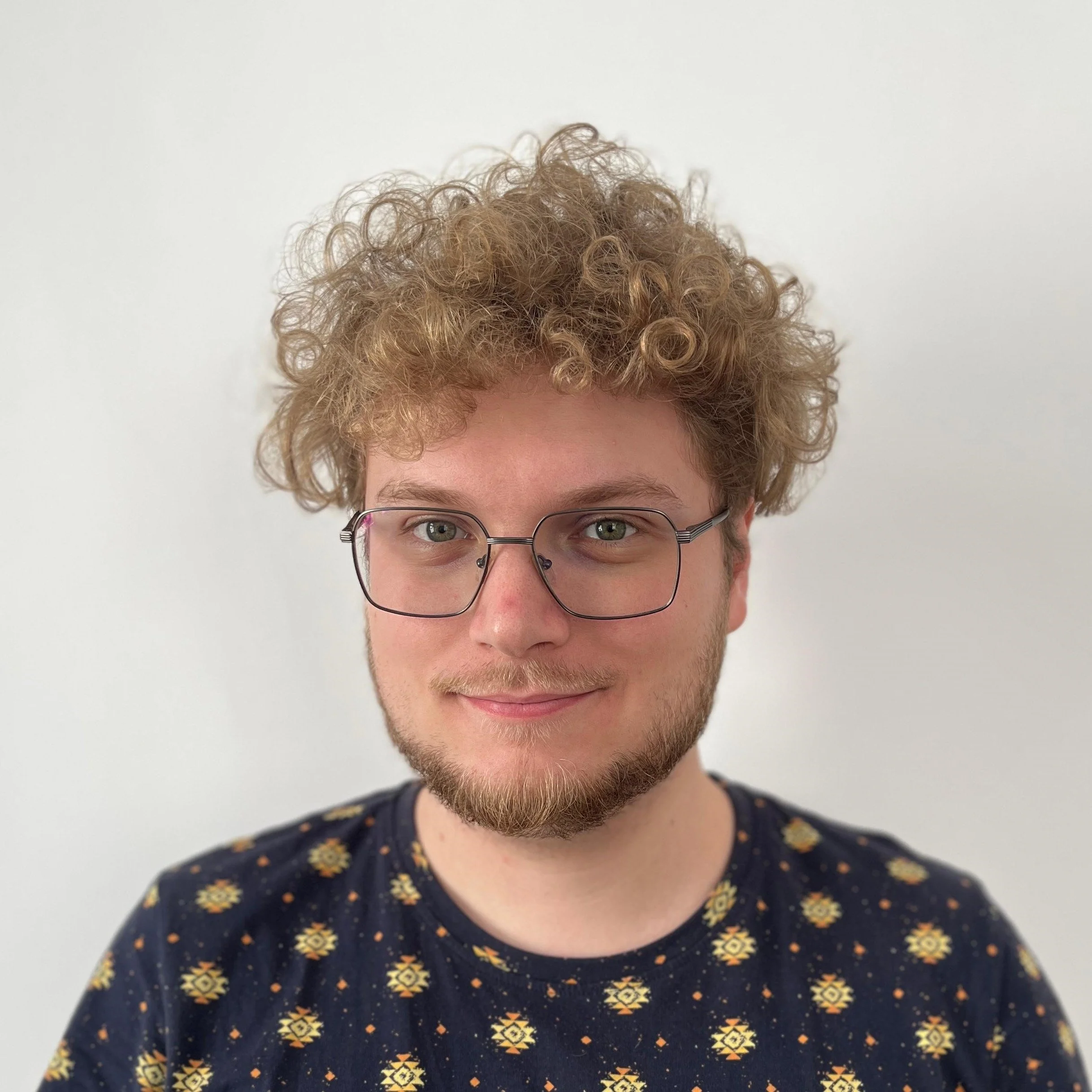 Portrait of a young man with curly blonde hair, glasses, and a light beard, smiling, wearing a dark shirt with a yellow floral pattern, against a plain white background.