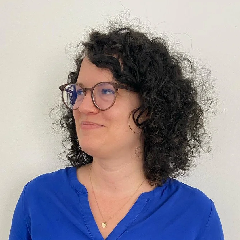 A woman with curly black hair, glasses, wearing a blue top and a gold necklace with a heart pendant, looking to the side against a plain light background.