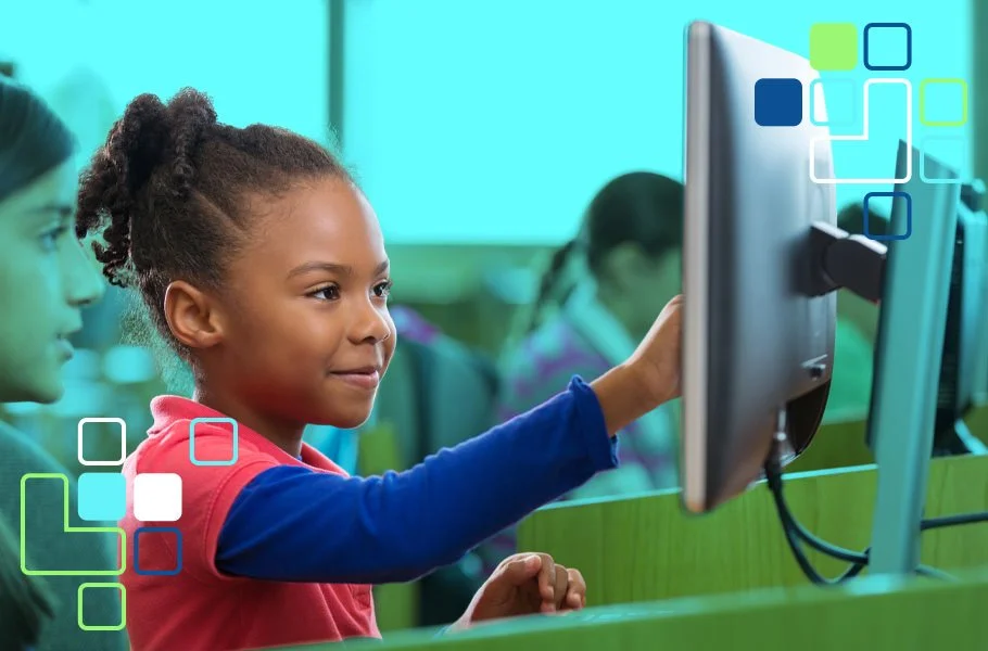 A young girl in a classroom using a computer, with other students in the background.