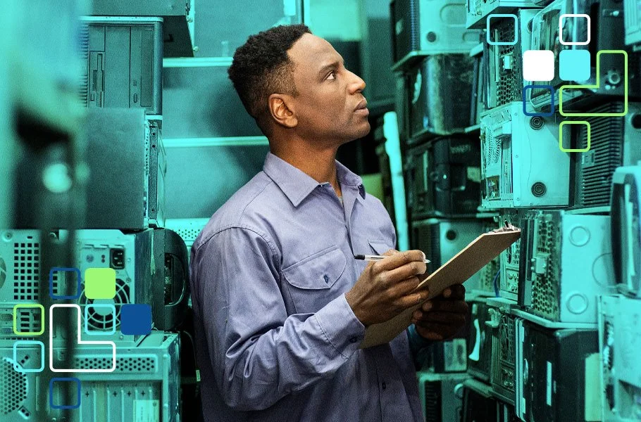 A man wearing a light purple shirt holding a clipboard and pen, standing among computer servers in a data center, inspecting the equipment.