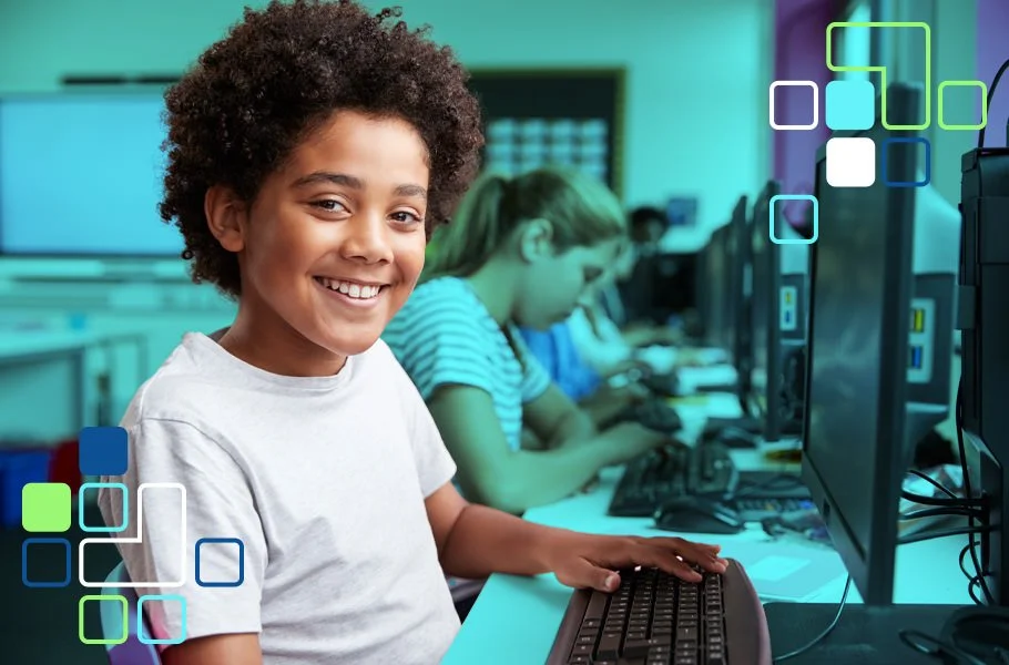 Smiling young boy in a computer classroom, sitting at a desk with a keyboard, with other children working on computers in the background.