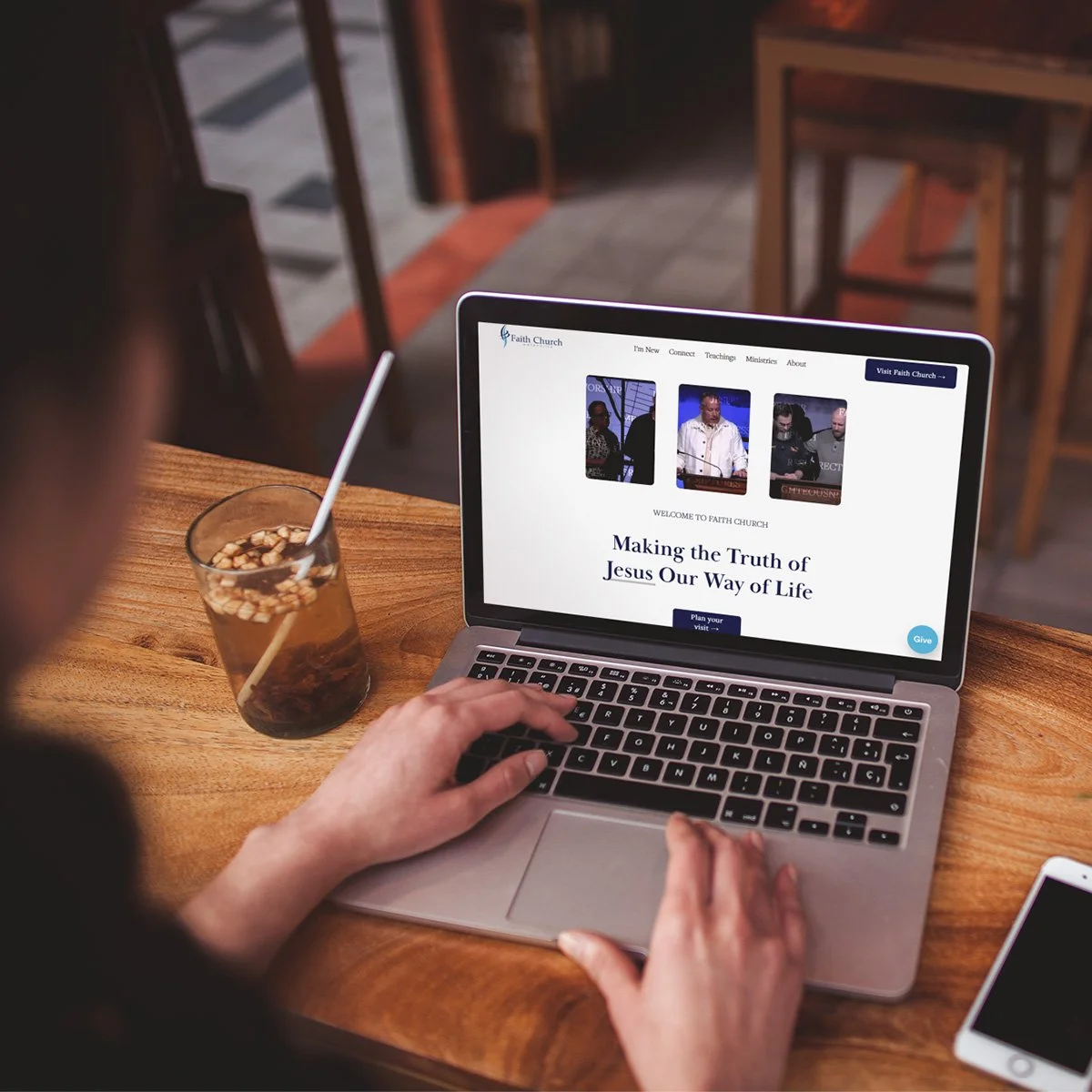 A person uses a laptop displaying a church website titled 'Making the Truth of Jesus Our Way of Life' at a wooden table with a glass of iced beverage and a straw, in a cozy cafe or restaurant setting.
