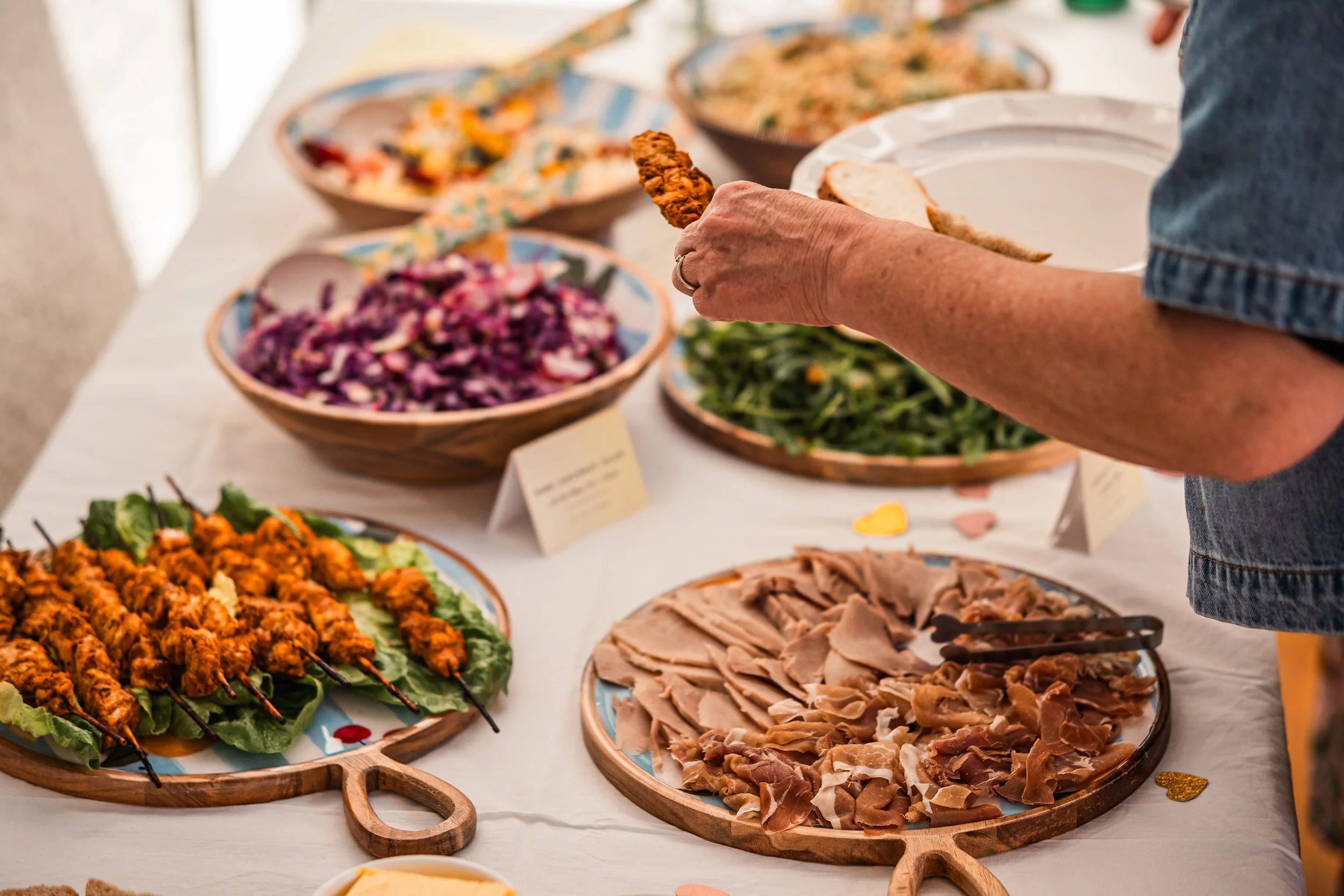Person serving themselves at a buffet with various dishes including sliced meat, skewered chicken, salad, and other colorful salads.
