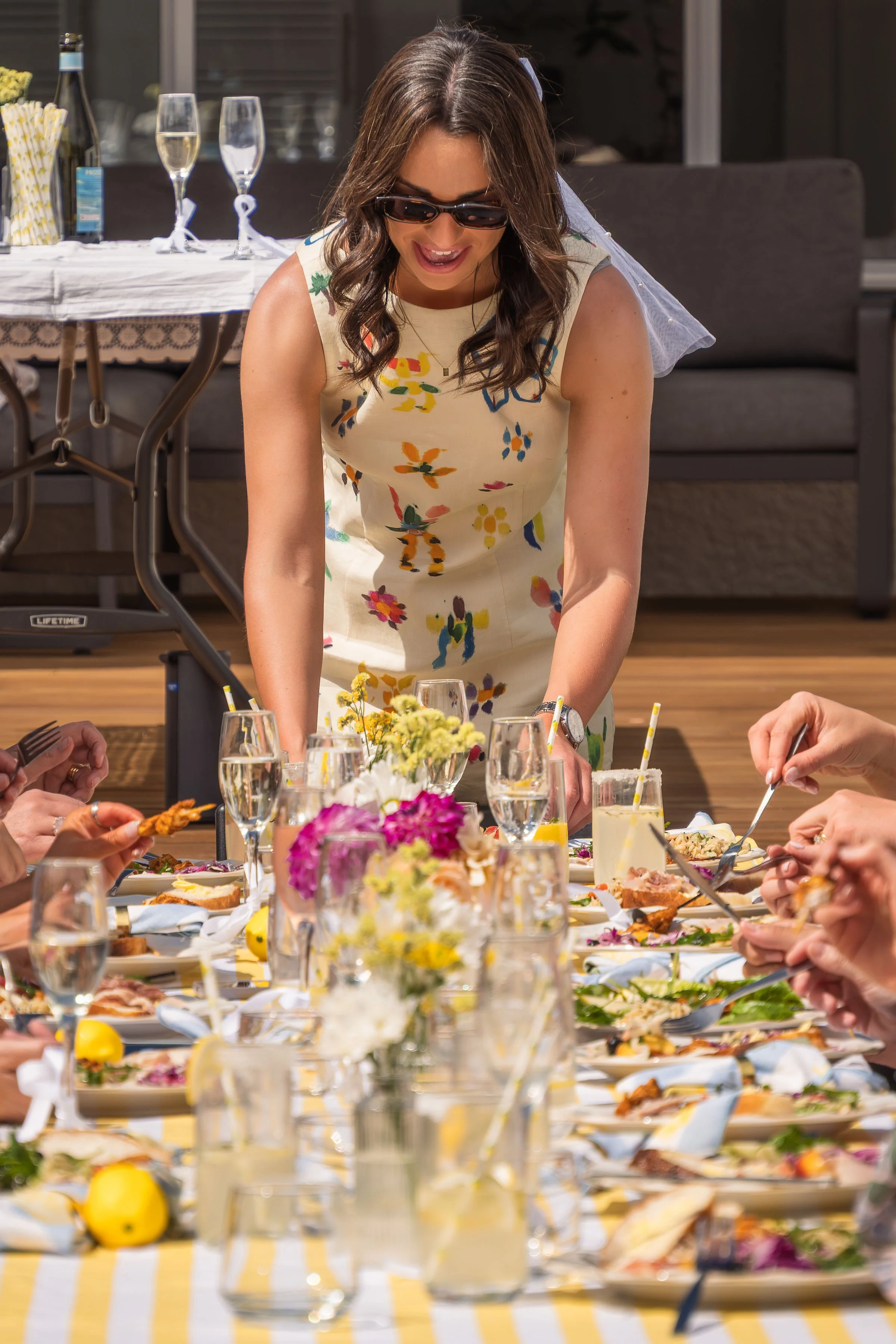 A woman wearing sunglasses and a floral dress is standing at a table, smiling and serving food at a gathering or celebration with several guests, food, drinks, and floral decorations on the table.