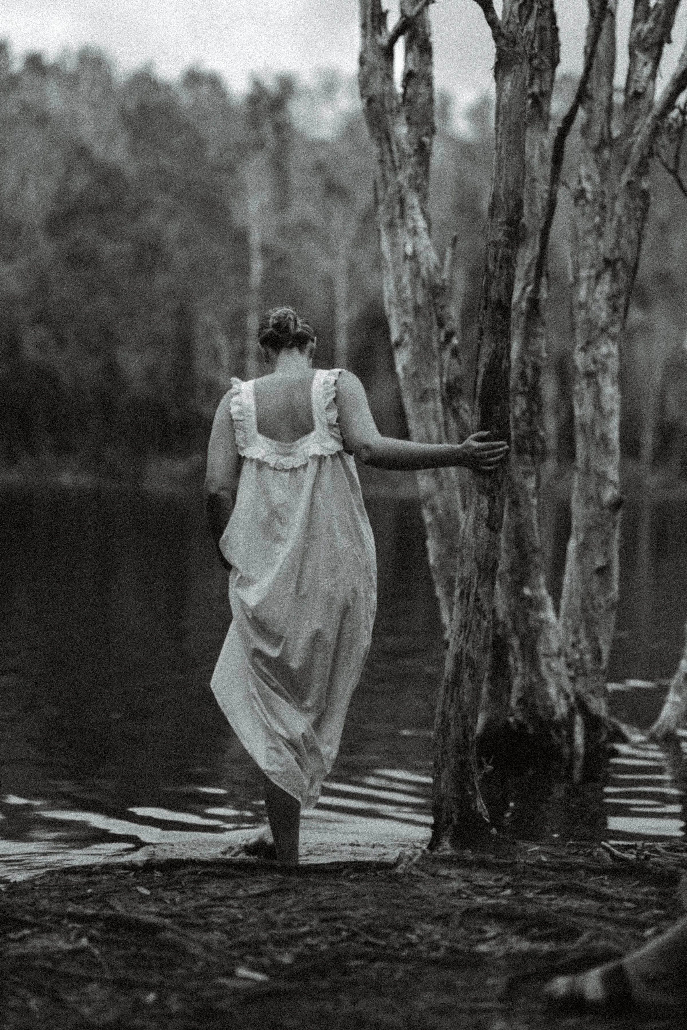 Woman in a long white dress walking barefoot through shallow water among paperbark trees