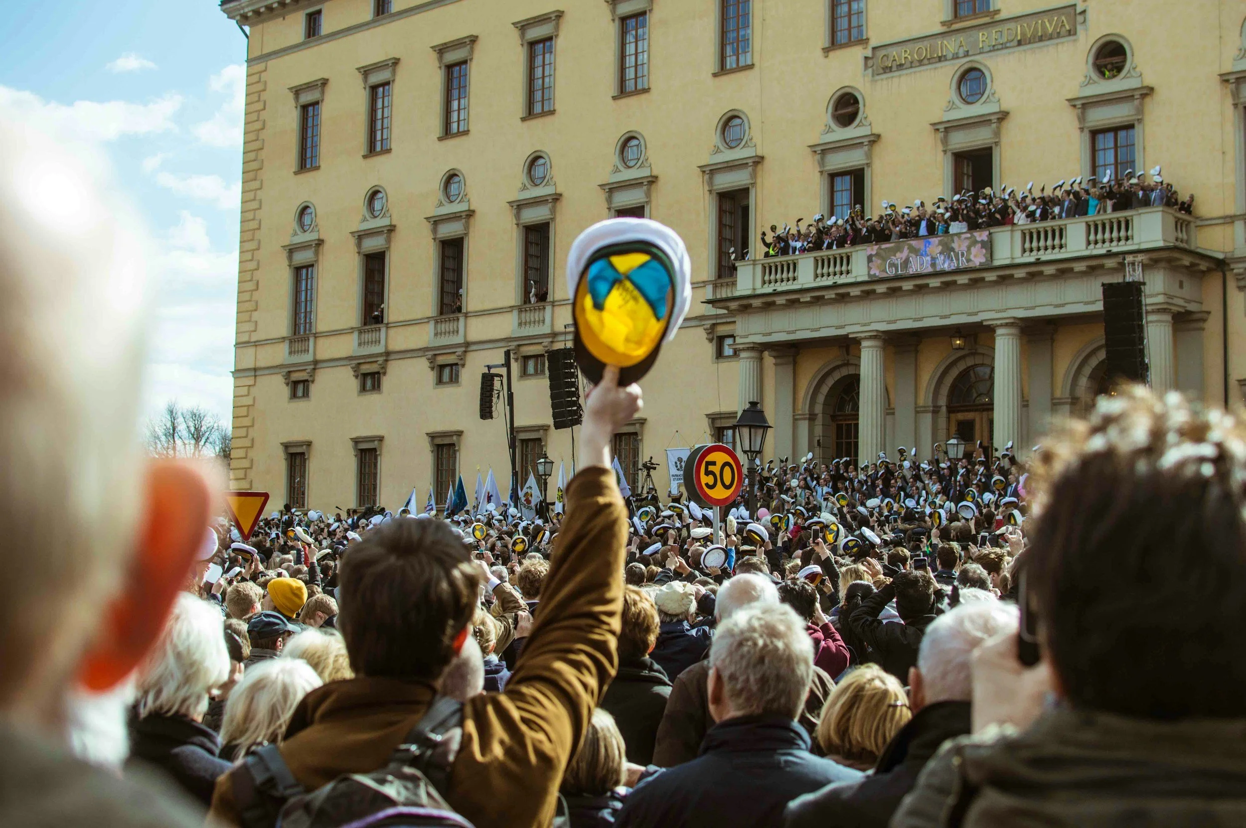 Folk samlat framför en byggnad för ett firande, många bär vita mössor med blå och gula detaljer, en person håller en mössa med en svensk flagga uppe, tydlig vår- eller sommaratmosfär.