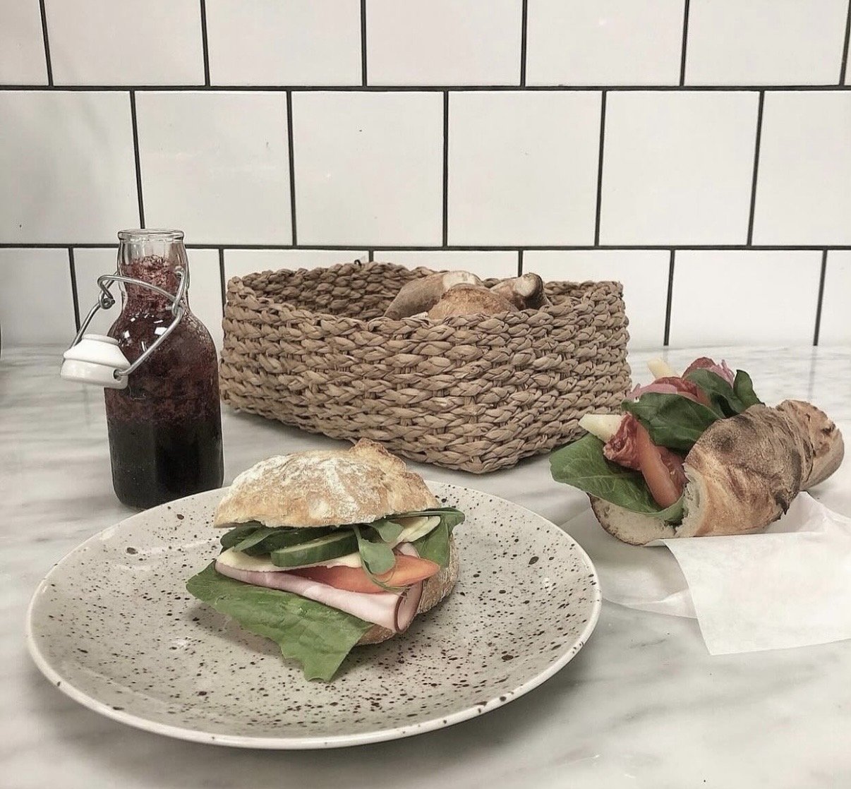 A sandwich with ham, tomato, cucumber, and leafy greens on a white speckled plate, a wrapped sandwich with similar contents, a basket with bread and mushrooms, and a small glass bottle of dark berry juice, all on a white marble table with white tiled wall background.