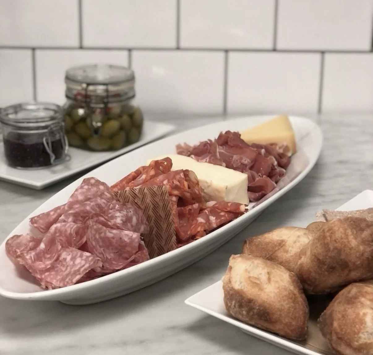 Ost, charkuterier och knötbröd på serveringsfat, med inlagda oliver och kryddor i bakgrunden.