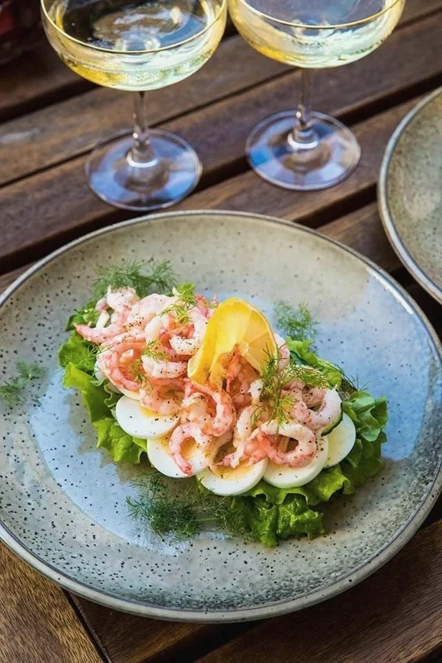 Sallad med räkor, hårdkokta äggskivor, en citronklyfta och dill på en grön salladsbas, serverad på en beige tallrik. Två glas vitt vin står ovanför tallriken på ett träbord.