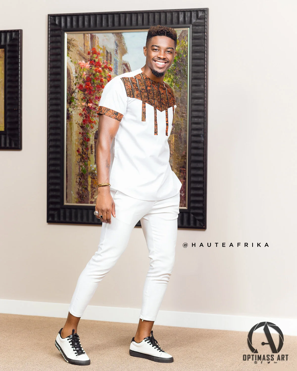 A smiling man in white pants and a white T-shirt with an African print detail, wearing sneakers, standing indoors in front of colorful artwork.