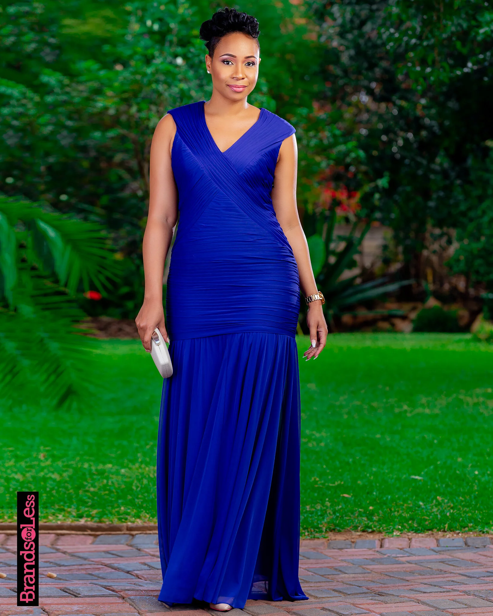 A woman in a long, royal blue gown standing outdoors on a brick pathway with green bushes and trees in the background.
