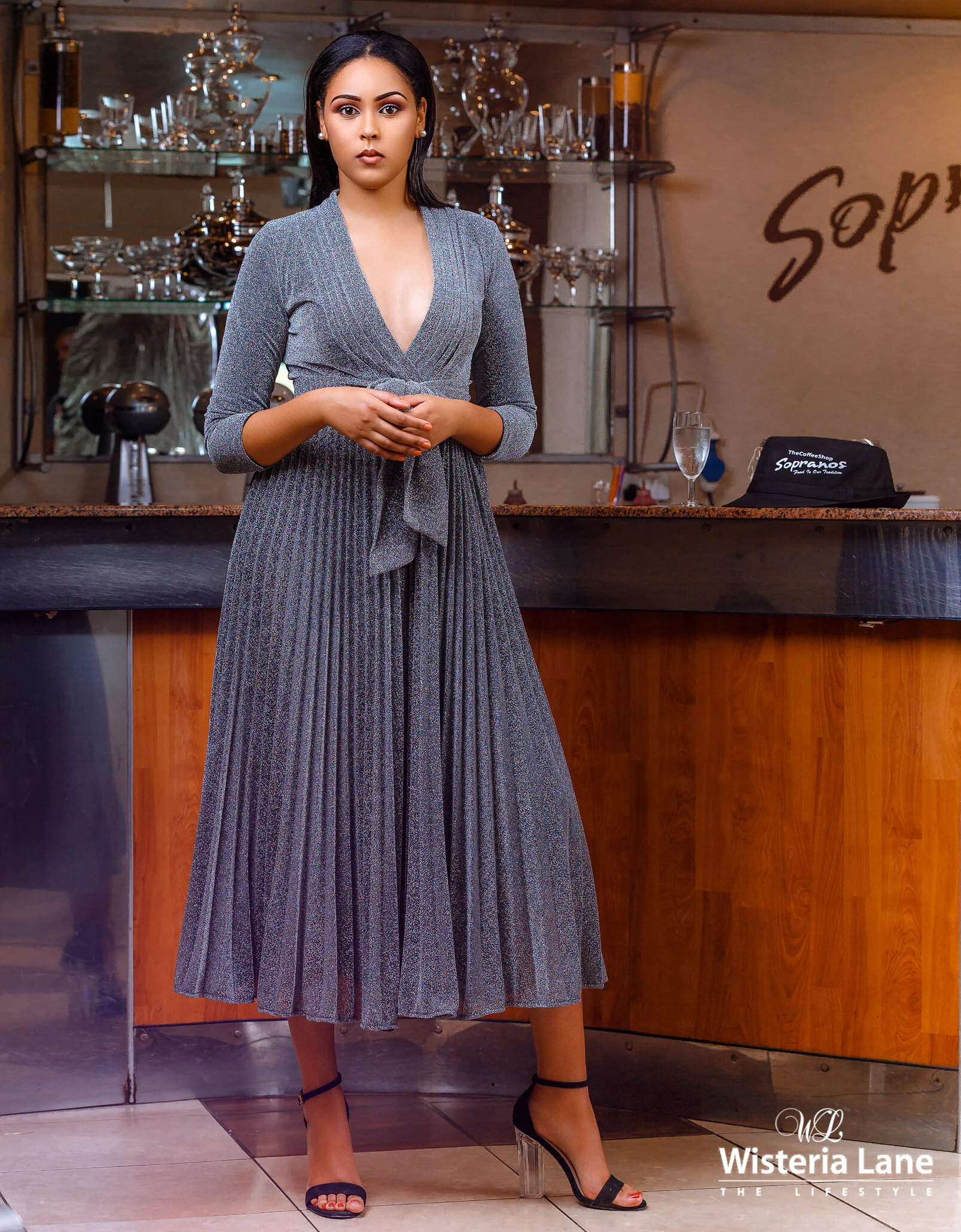A woman stands at a bar counter in a fashionable outfit, with a glass of champagne beside her. The background includes glassware and a partially visible sign that reads 'Sopras.' She wears a long, pleated gray dress with a deep V neckline, and black 