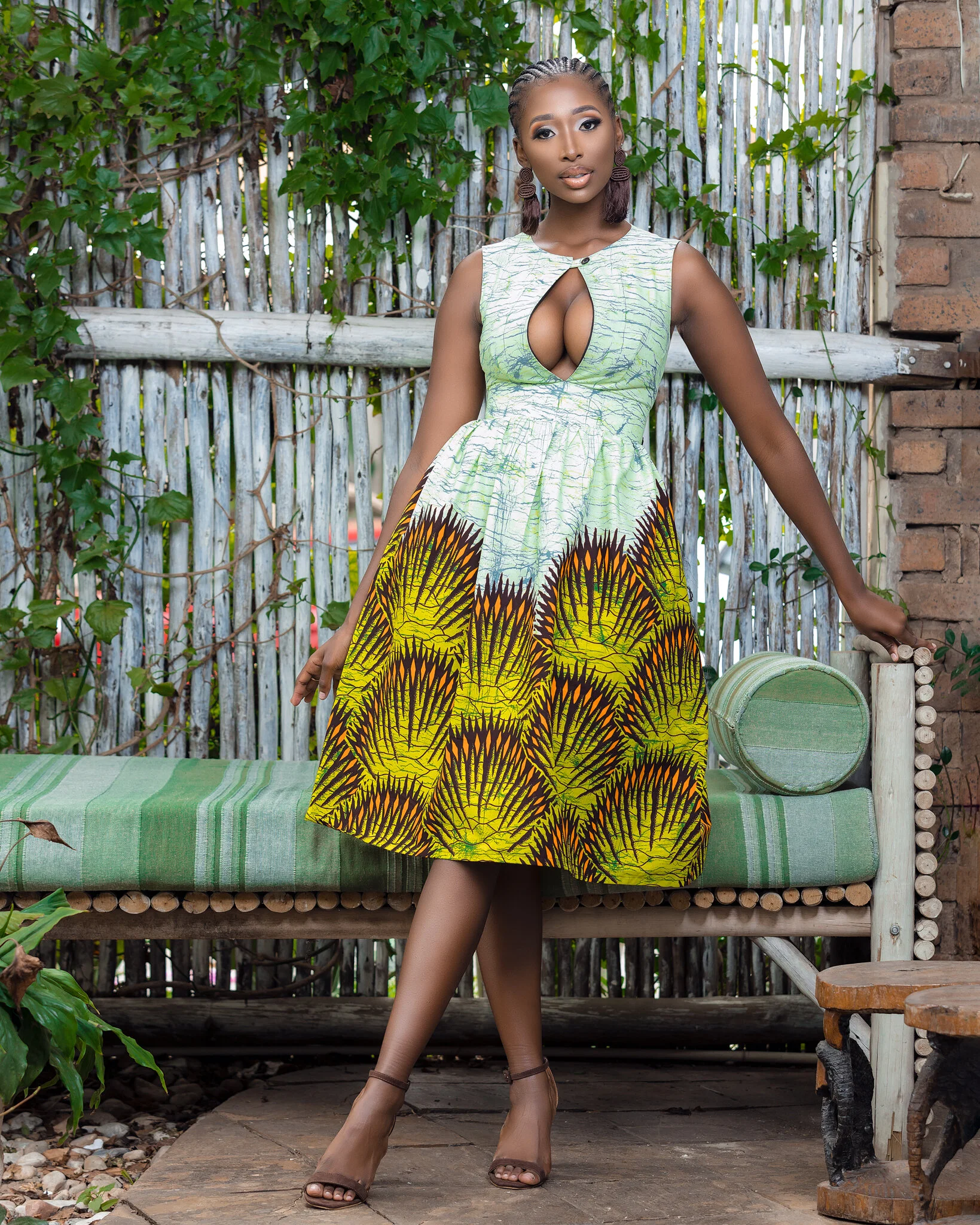 A woman standing outdoors in front of a wooden fence and lush green ivy, wearing a sleeveless, patterned dress with a keyhole cutout at the chest.
