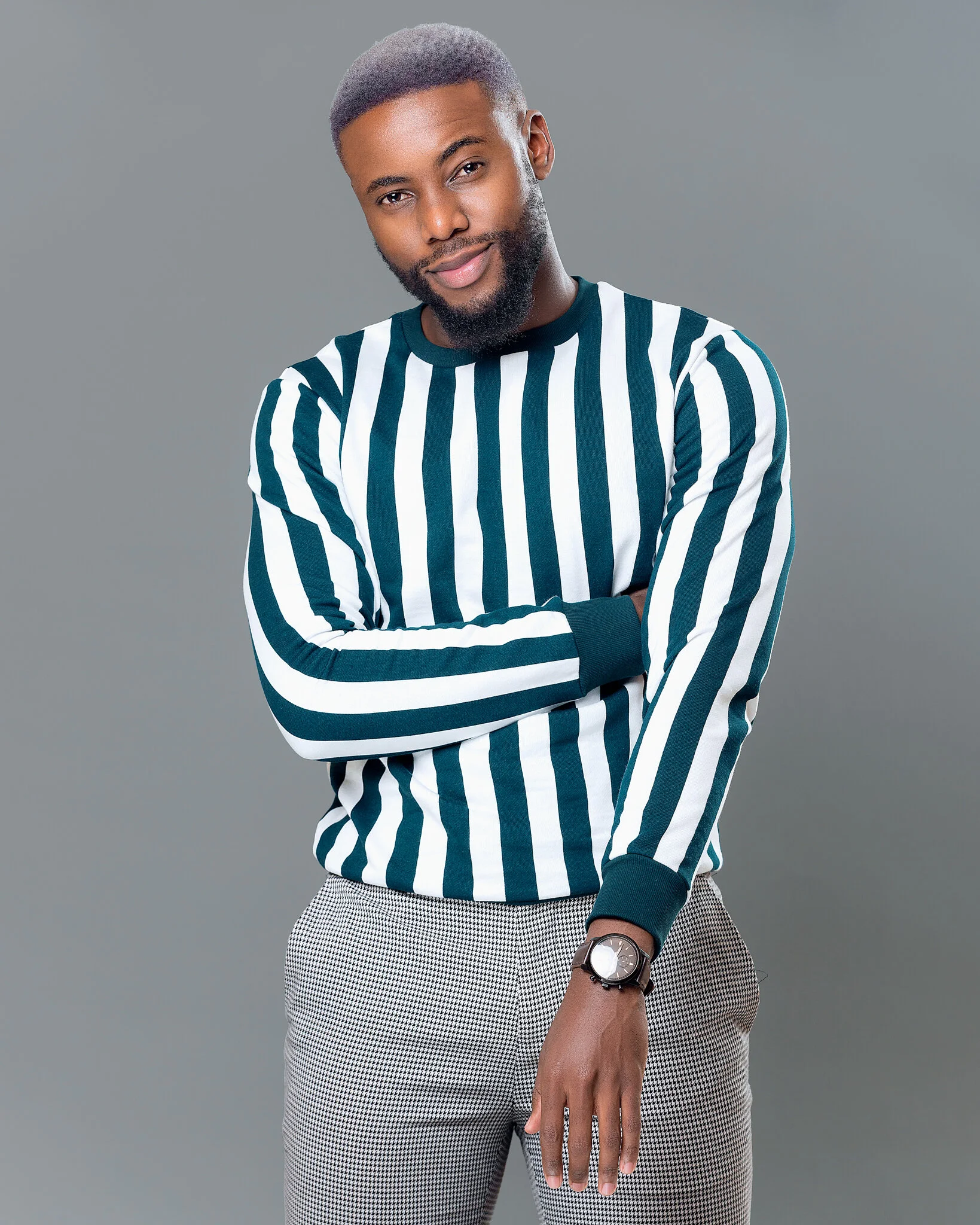 A young man with a beard and short hair, wearing a striped long-sleeve shirt and light-colored checkered pants, standing with arms crossed and smiling at the camera against a gray background.