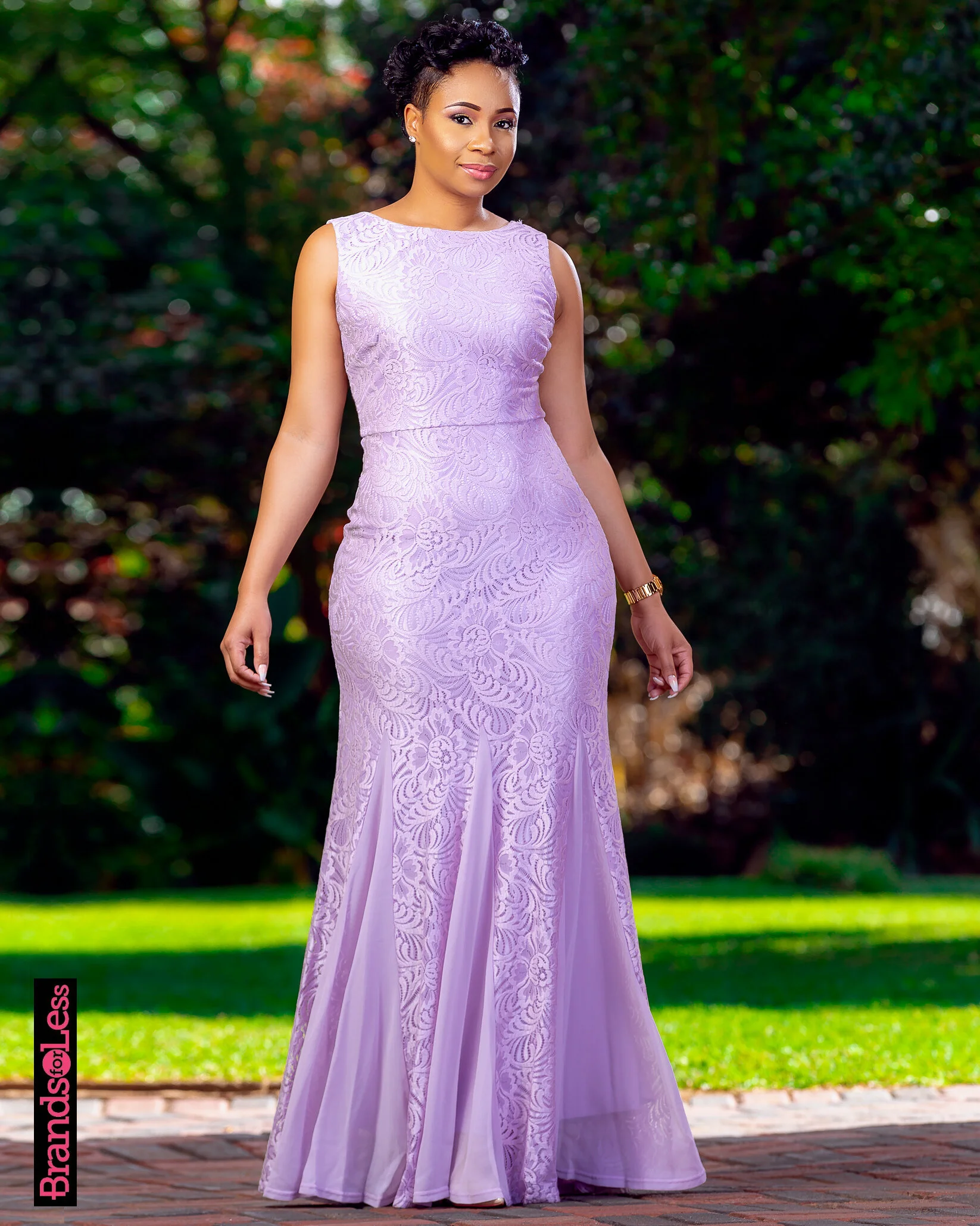 Woman in a long, lilac-colored, lace dress standing outdoors on a brick pathway with green trees in the background.