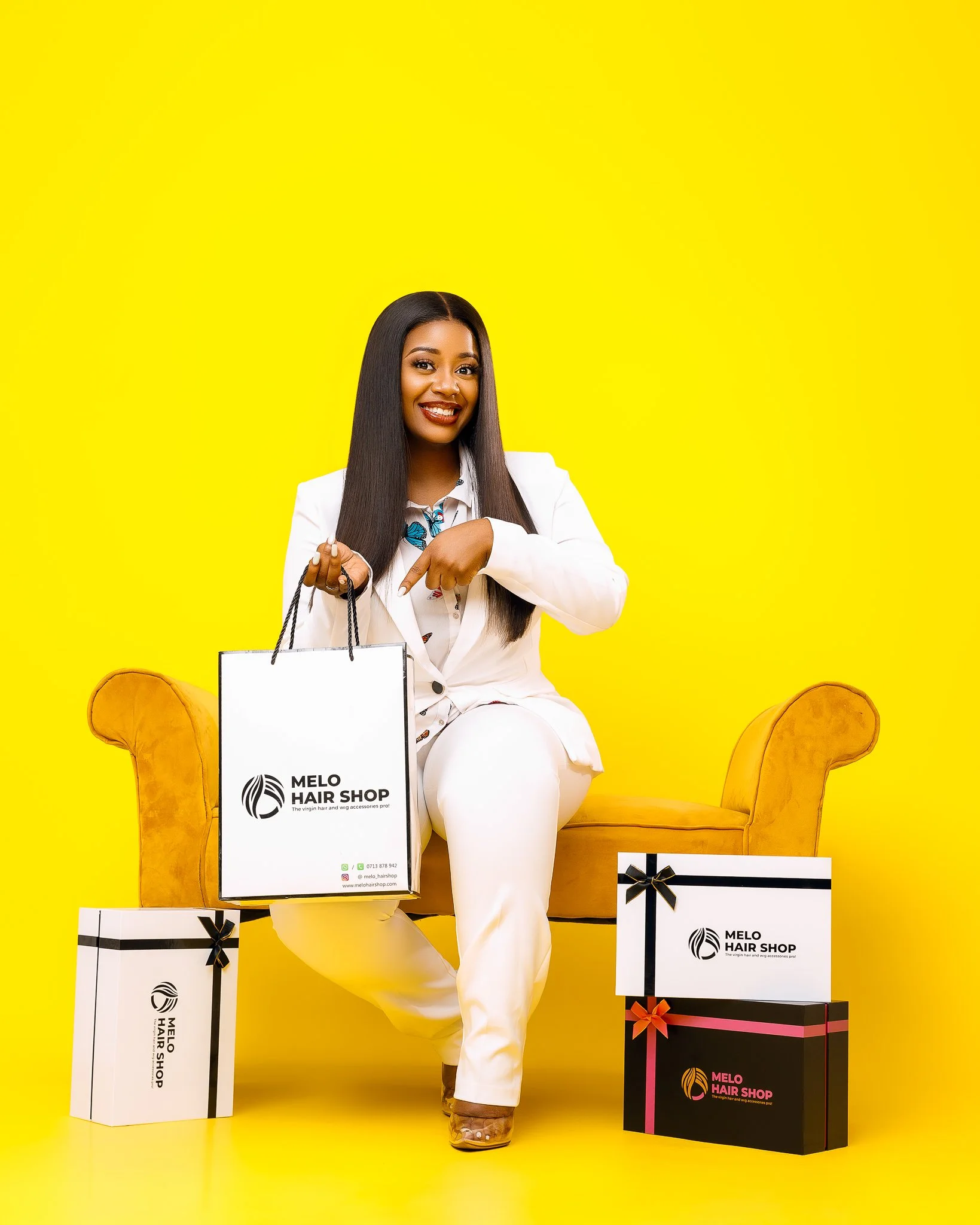 A woman with long, straight black hair, wearing a white suit, smiling and sitting on a yellow sofa. She is holding a shopping bag labeled 'Melo Hair Shop' with a logo. There are two large gift boxes, one white with black ribbon, and one black with pi