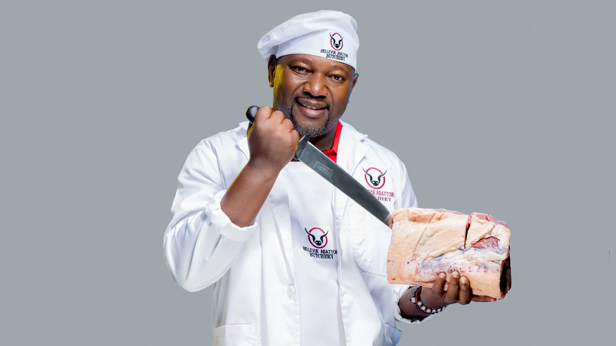 A man in a white chef uniform and chef hat, with a logo that reads "Bellevue Abattoir Butchery," is holding a large piece of meat and a large knife.