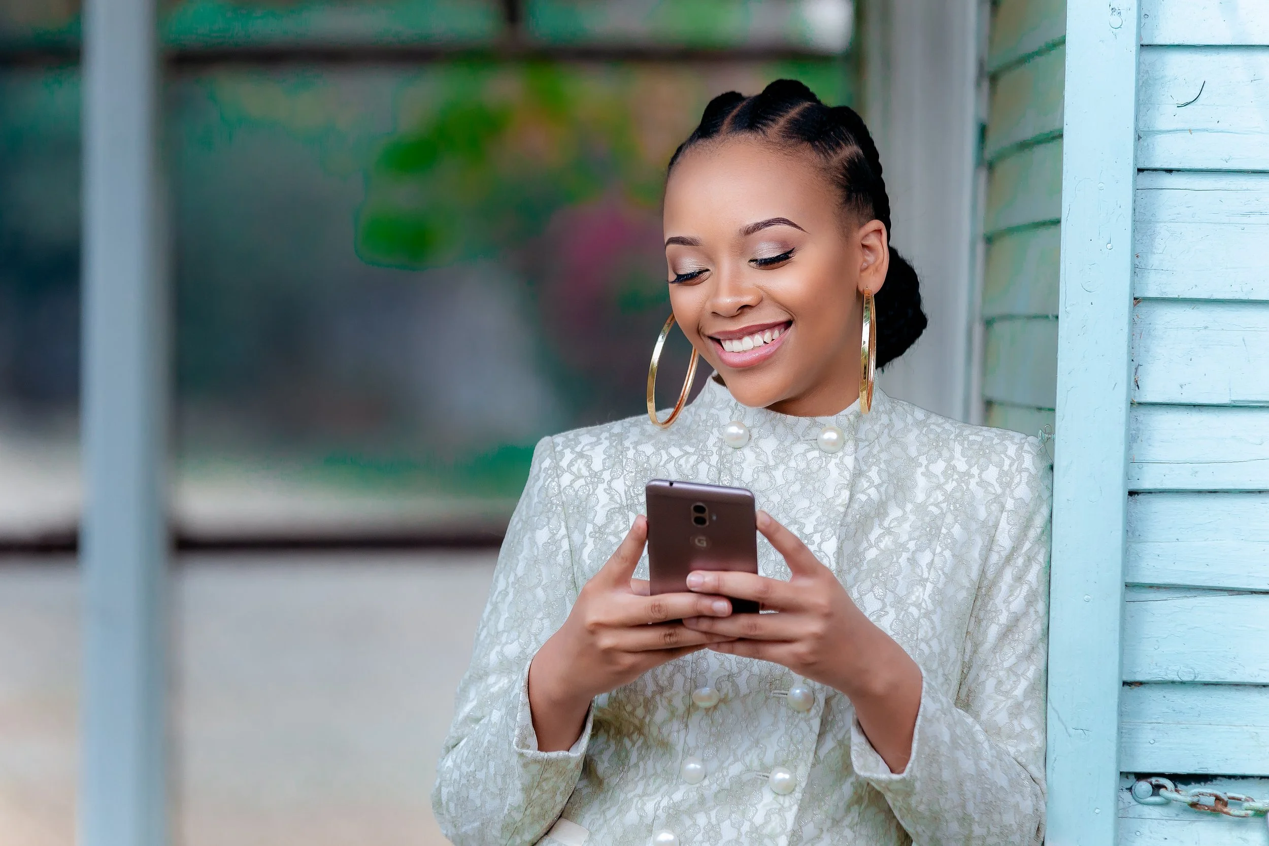 A woman with braided hair wearing large hoop earrings and a cream-colored, textured jacket with pearl buttons, smiling while looking at her smartphone outdoors near a light blue wooden structure.